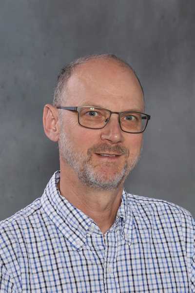 Ein lächelnder Mann mit Brille, der ein kariertes Hemd trägt und einen grauen Hintergrund hat.