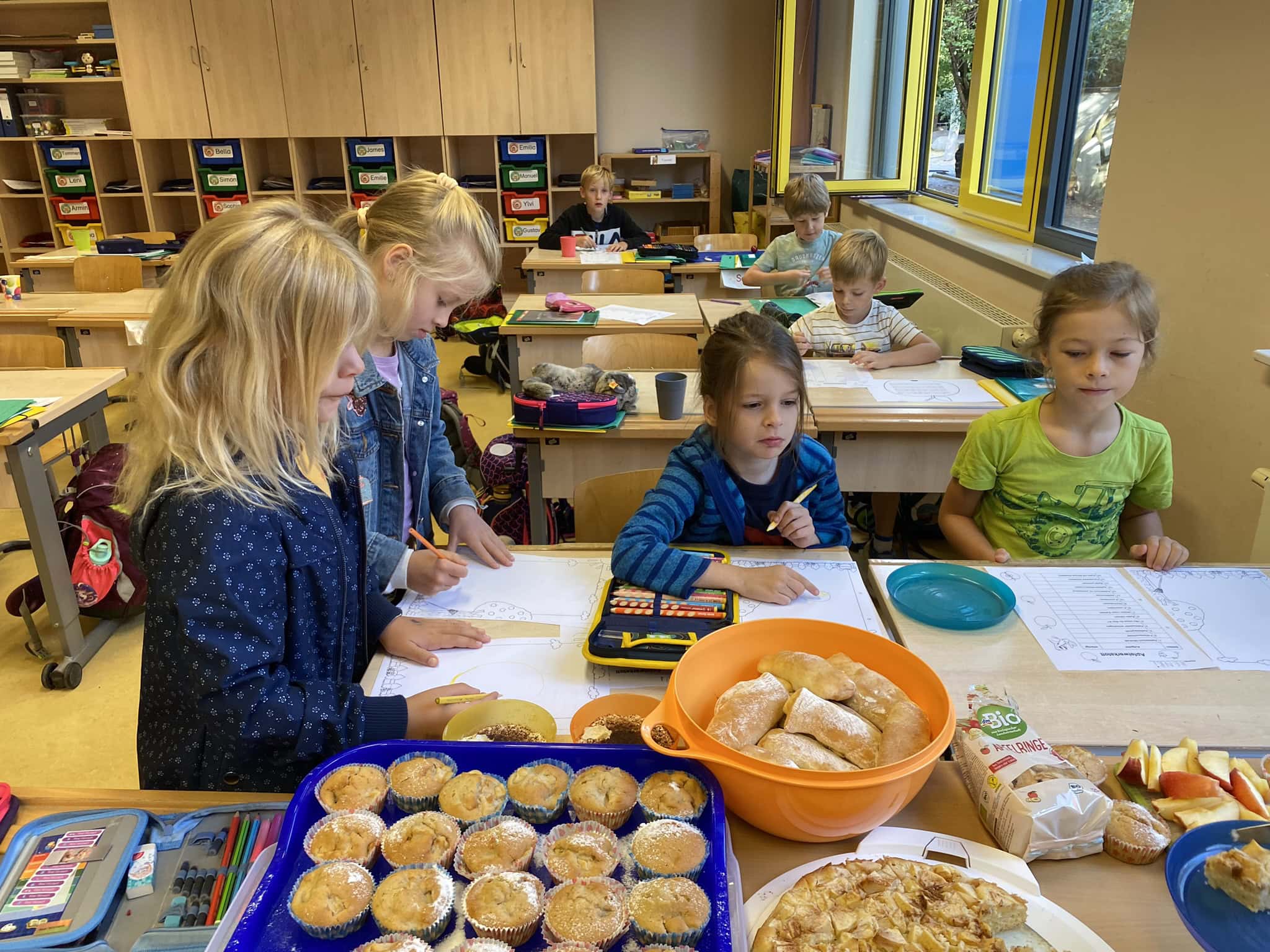 Vier Kinder in einem Klassenzimmer, freudig am Zeichnen, mit Gebäck auf den Tischen.