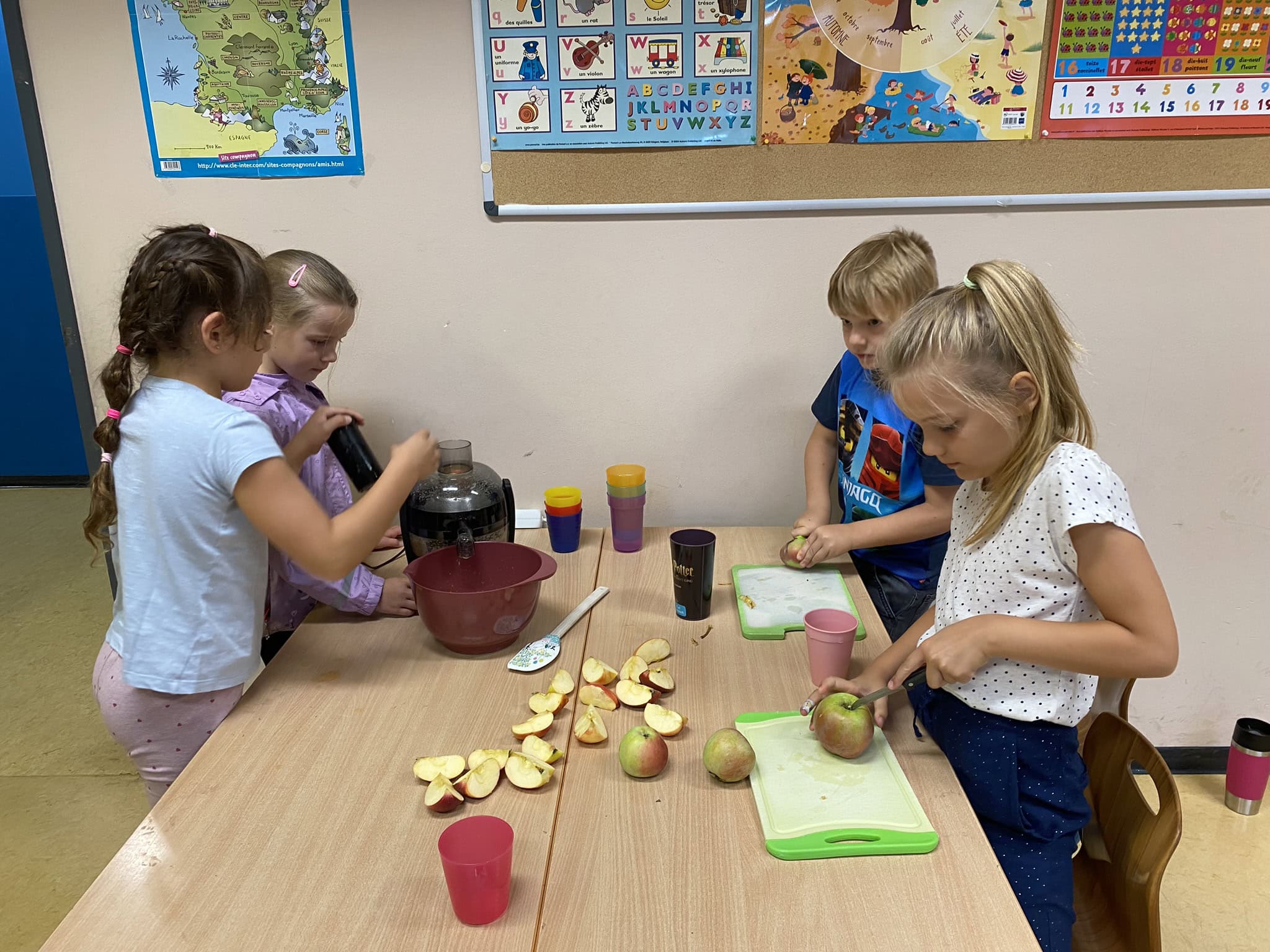 Vier Kinder lächeln und schneiden Äpfel in einem Klassenzimmer; an einer Tafel im Hintergrund.