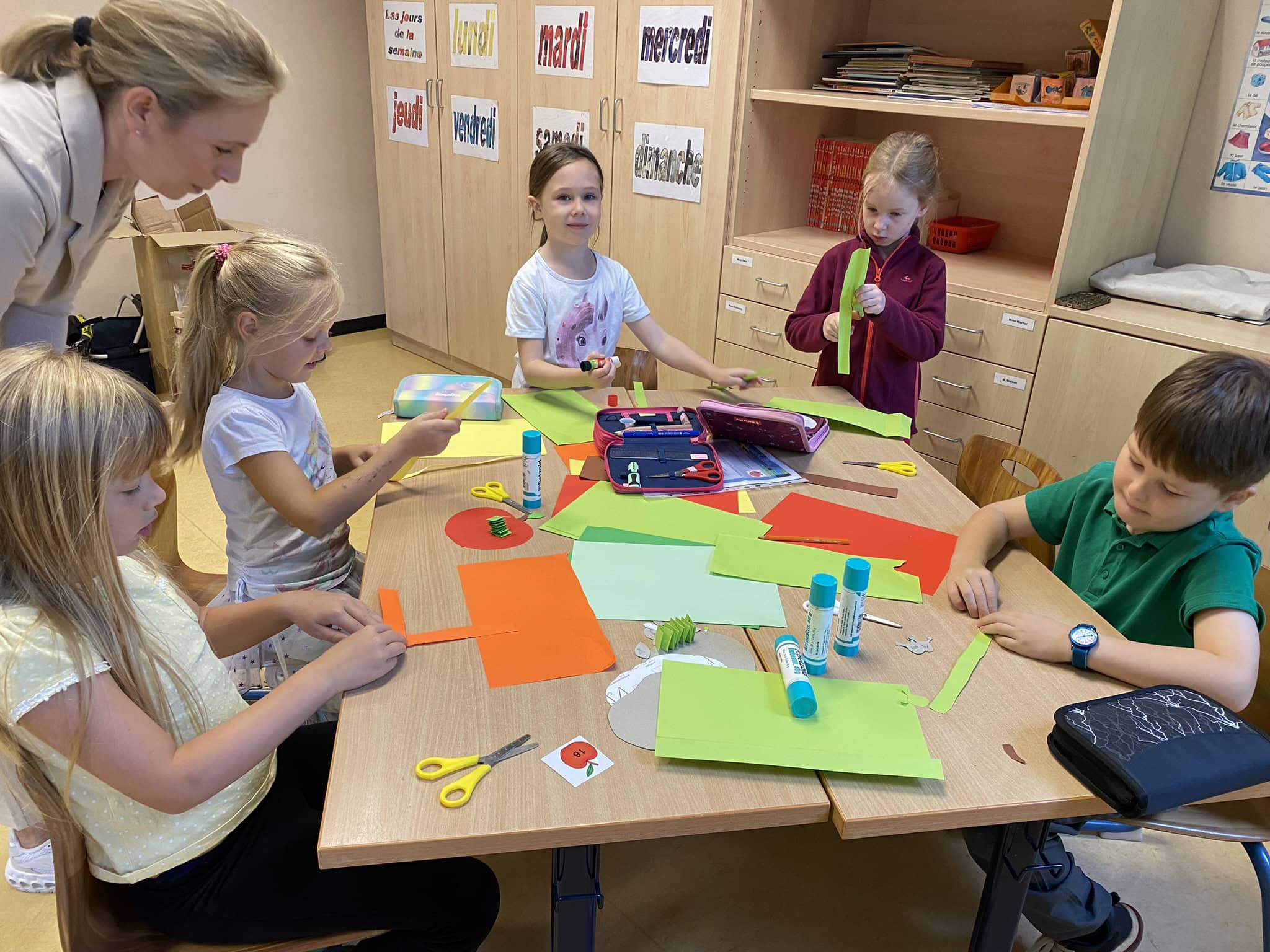 Kinder basteln konzentriert mit buntem Papier im Klassenzimmer. Lehrerin schaut zu.