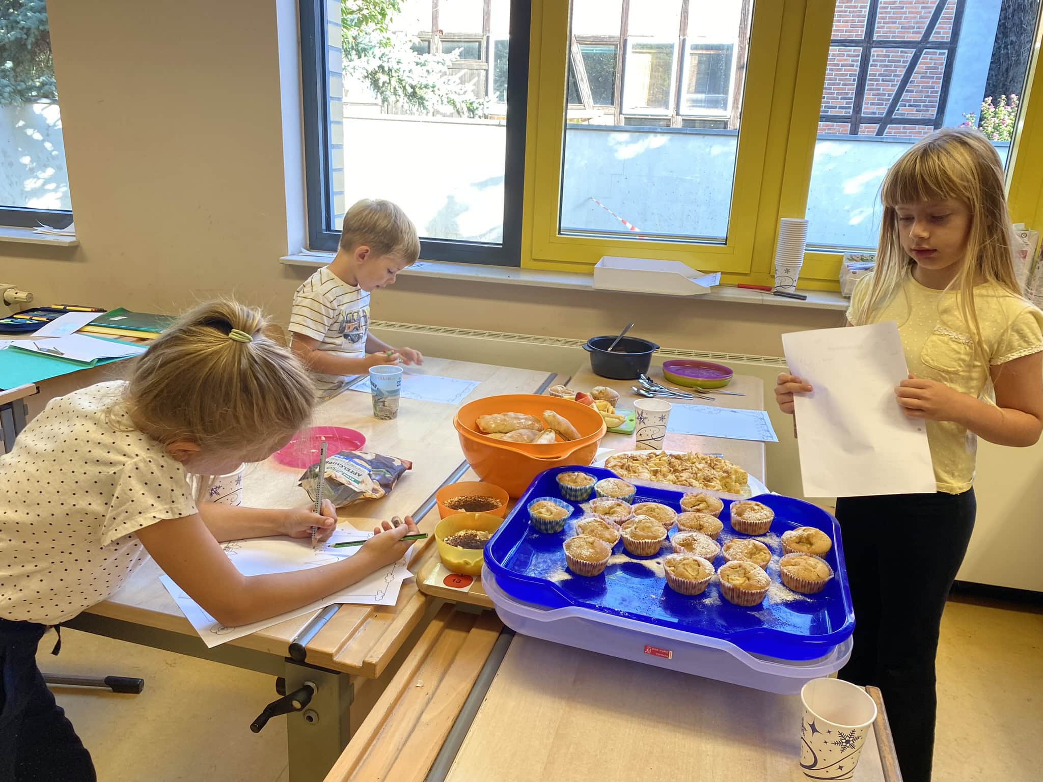 Drei Kinder in einem Klassenzimmer mit Backwaren und Bastelmaterialien, fröhlich beschäftigt.