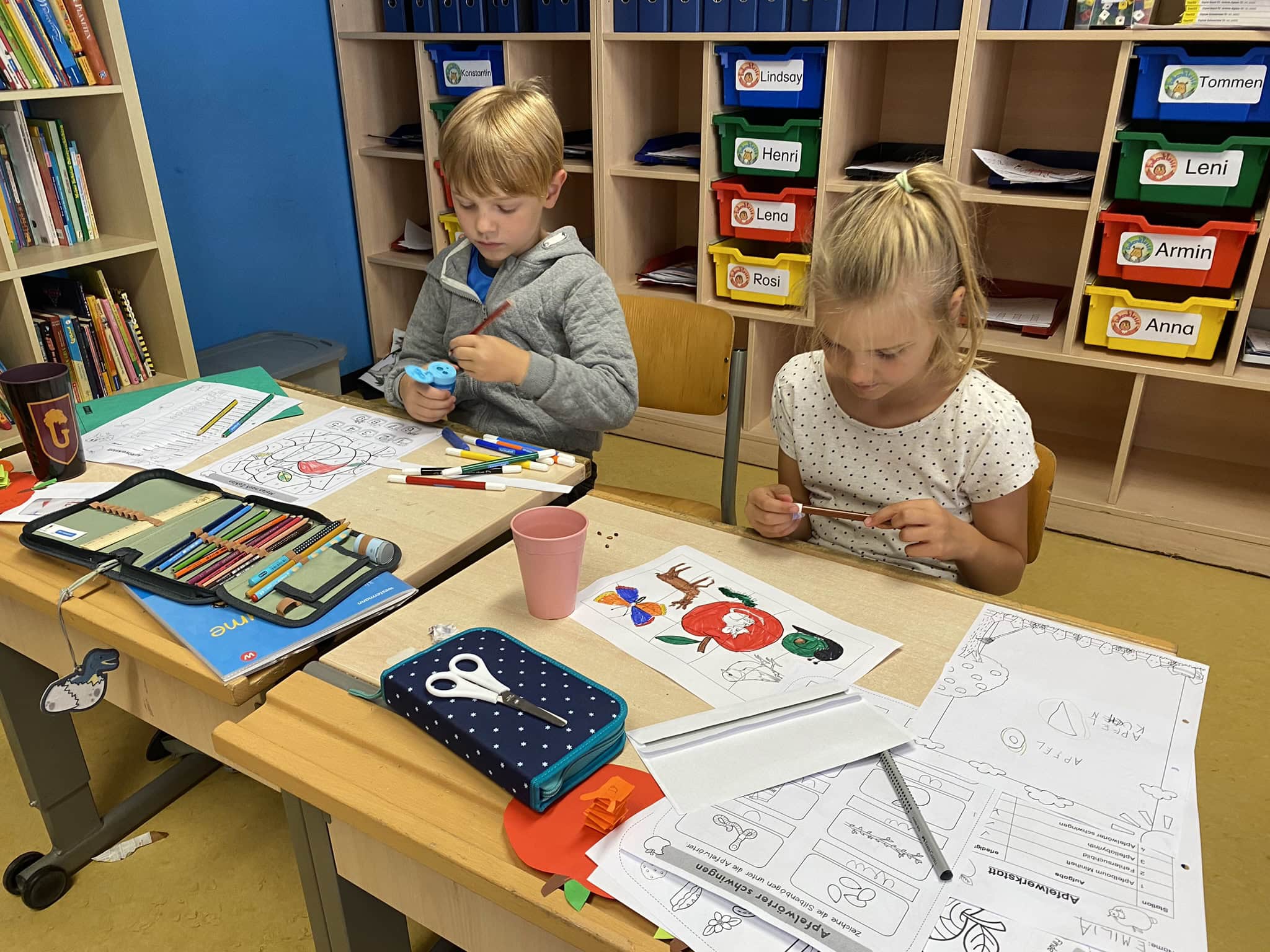 Zwei Kinder konzentriert am Basteln; farbenfrohe Stifte und gestaltetes Papier auf dem Tisch.