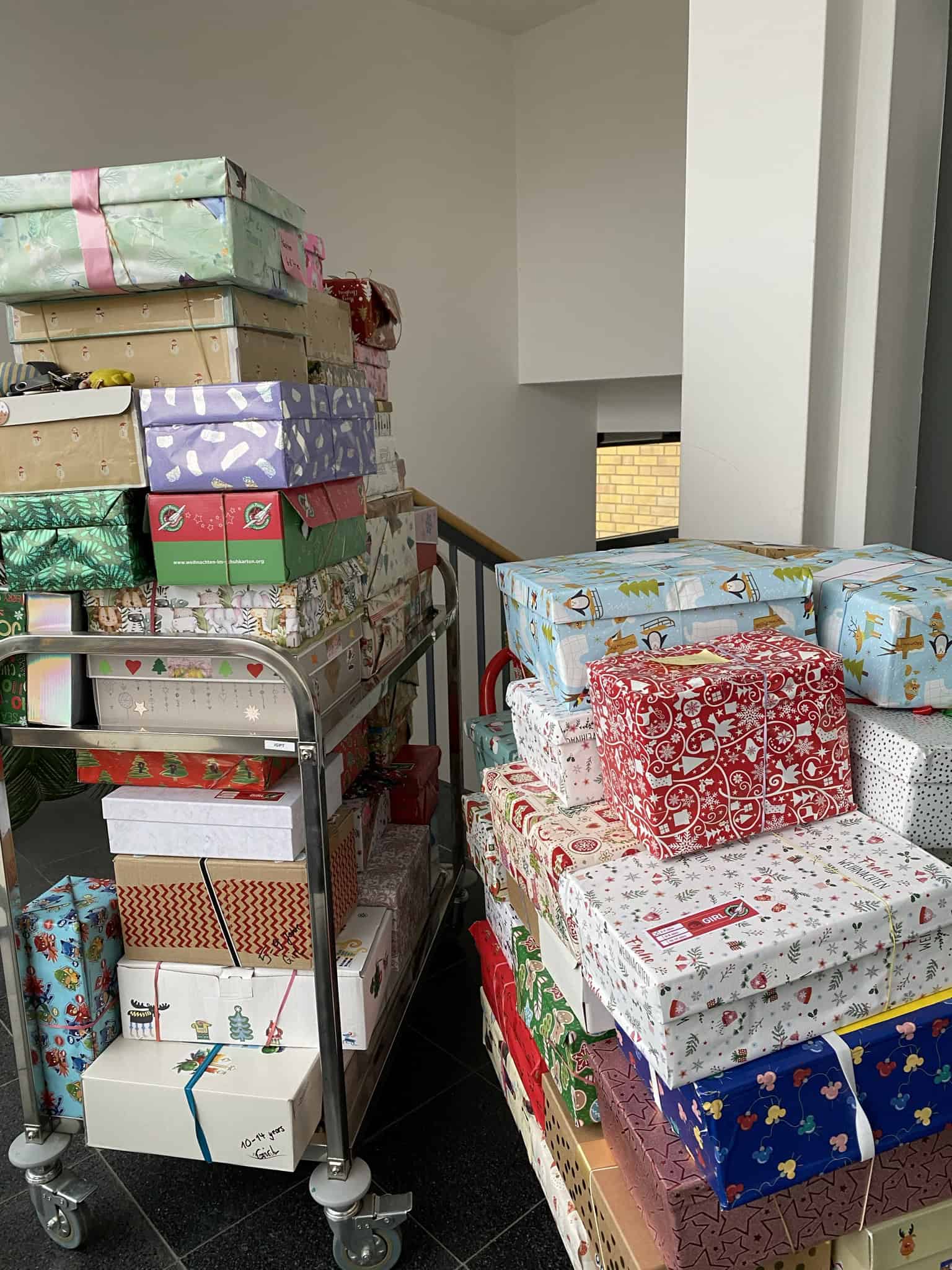 Zwei Stapel von bunt verpackten Geschenken auf einem Rollwagen in einem Treppenhaus.