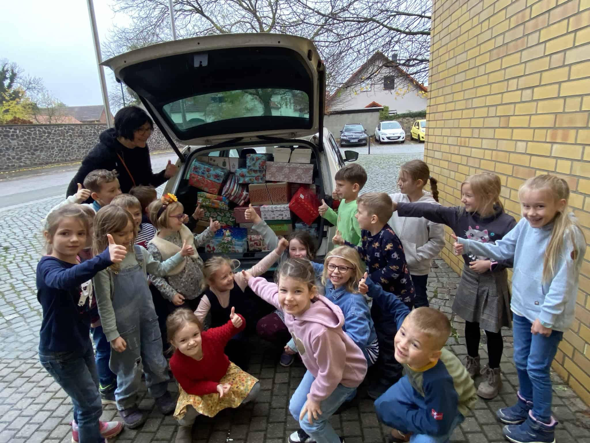 Eine Gruppe fröhlicher Kinder mit Geschenken vor einem Auto mit offenem Kofferraum.