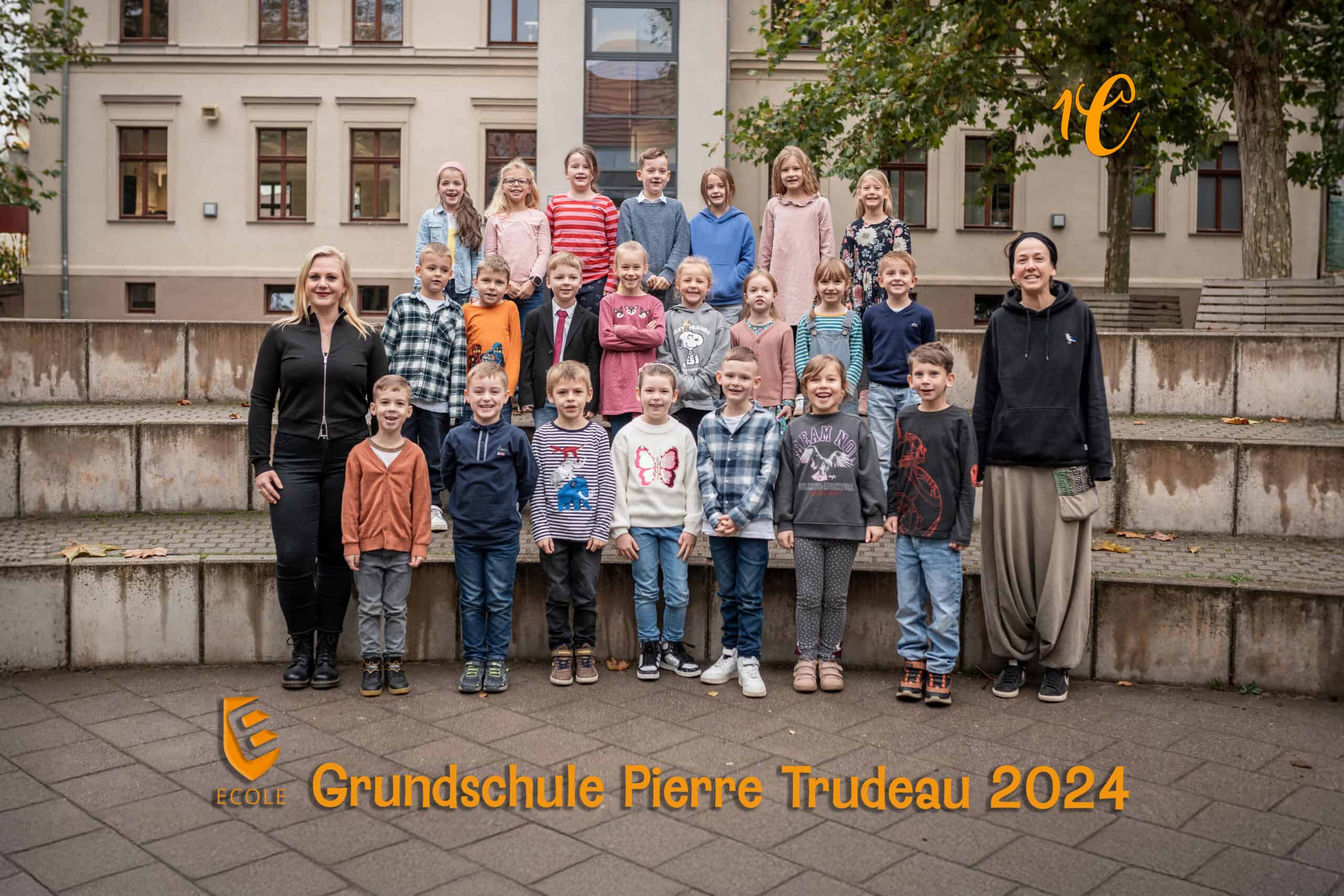Eine Gruppe von 23 Personen lächelt draußen, Kleidung in bunten Farben, Schulgebäude im Hintergrund.