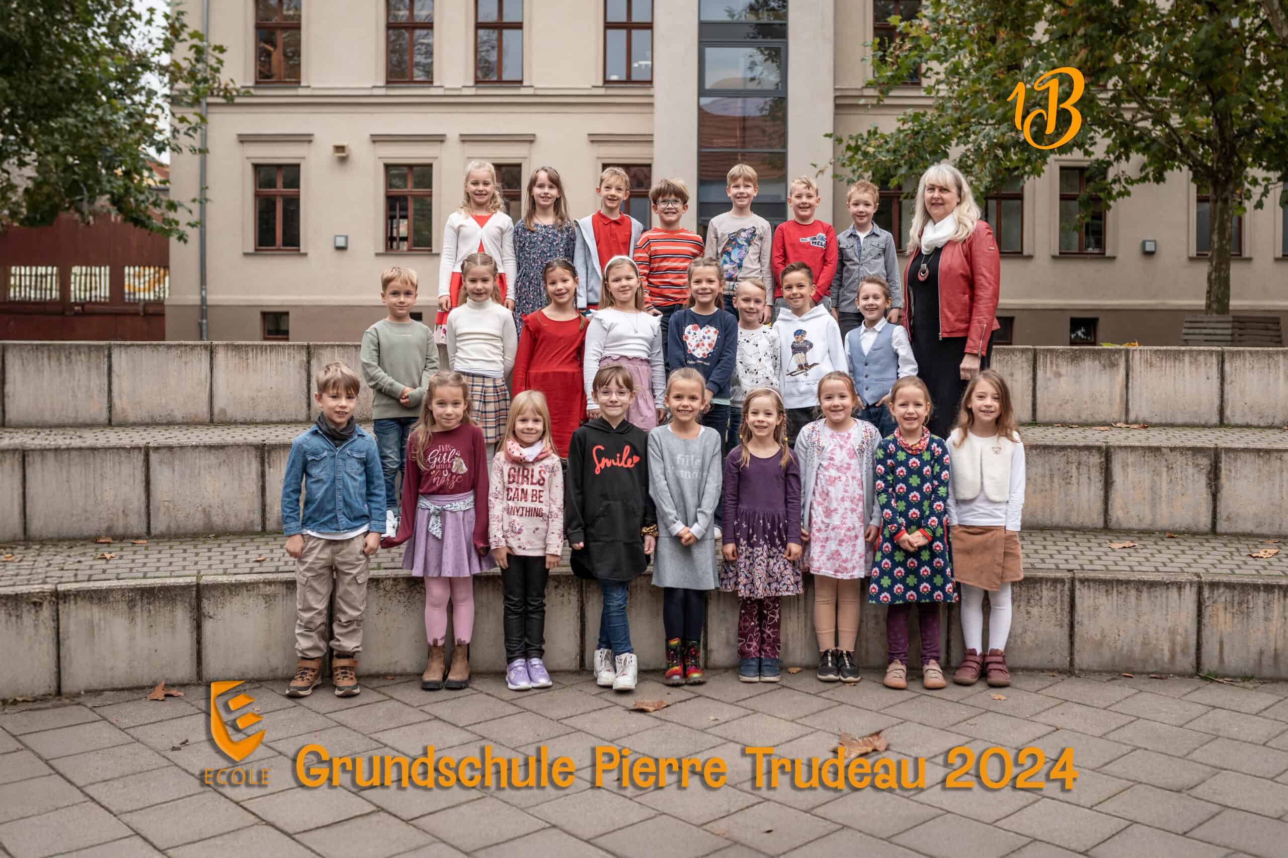 Schulklasse mit Lehrerin draußen, Kinder lächeln, bunte Kleidung, herbstliche Umgebung.