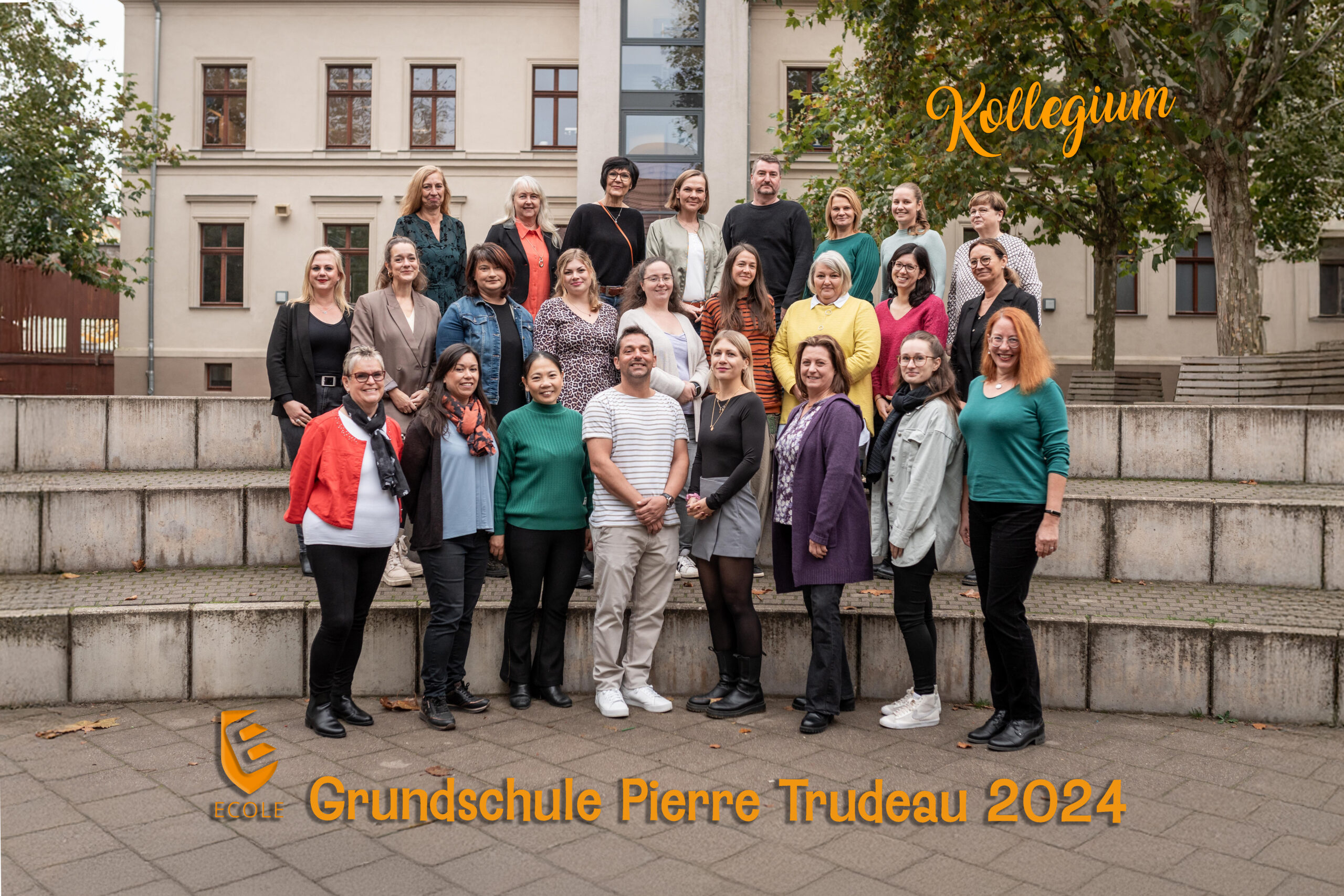 Gruppe von 25 lächelnden Menschen draußen, Herbstfarben, Schulgebäude im Hintergrund.