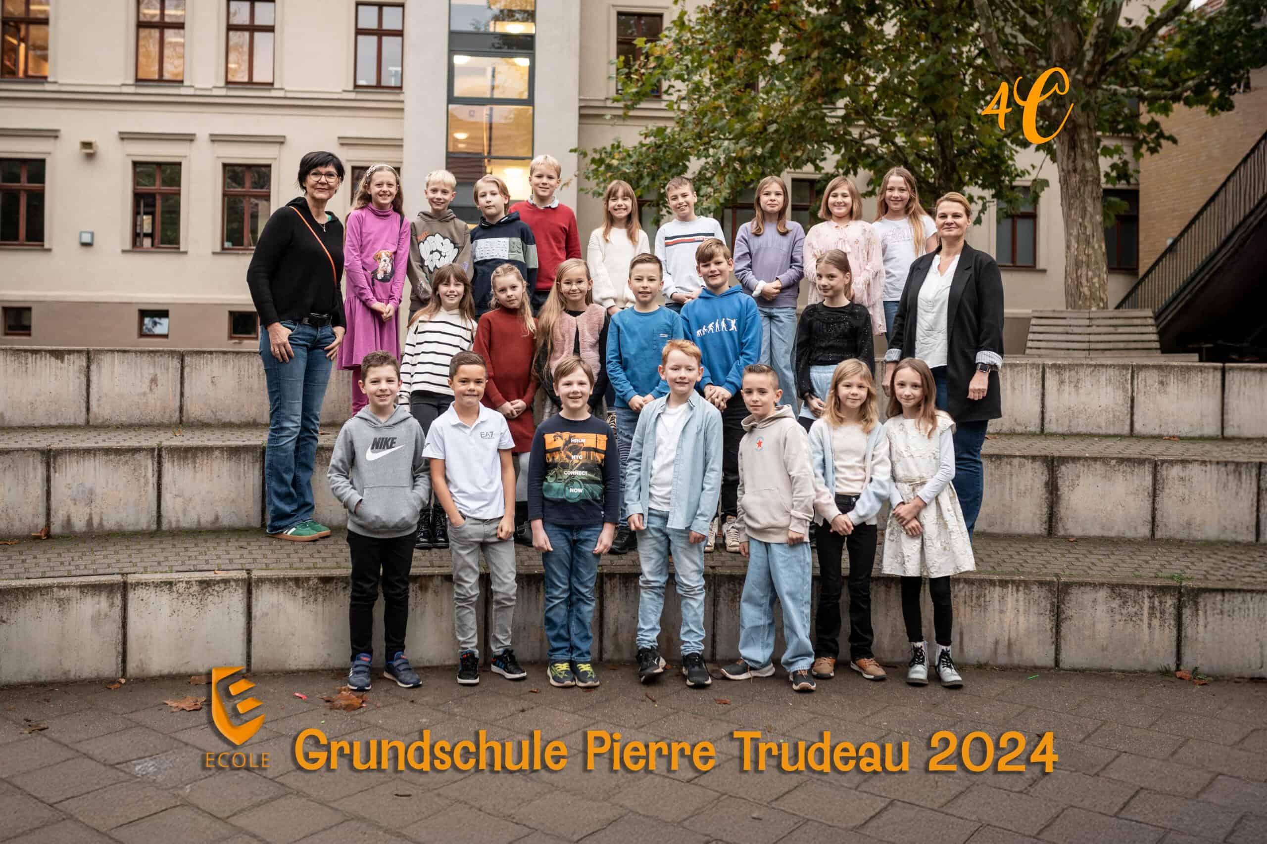Gruppe von Kindern und zwei Erwachsenen auf Stufen vor einem Schulgebäude, lächeln, herbstliche Kleidung.