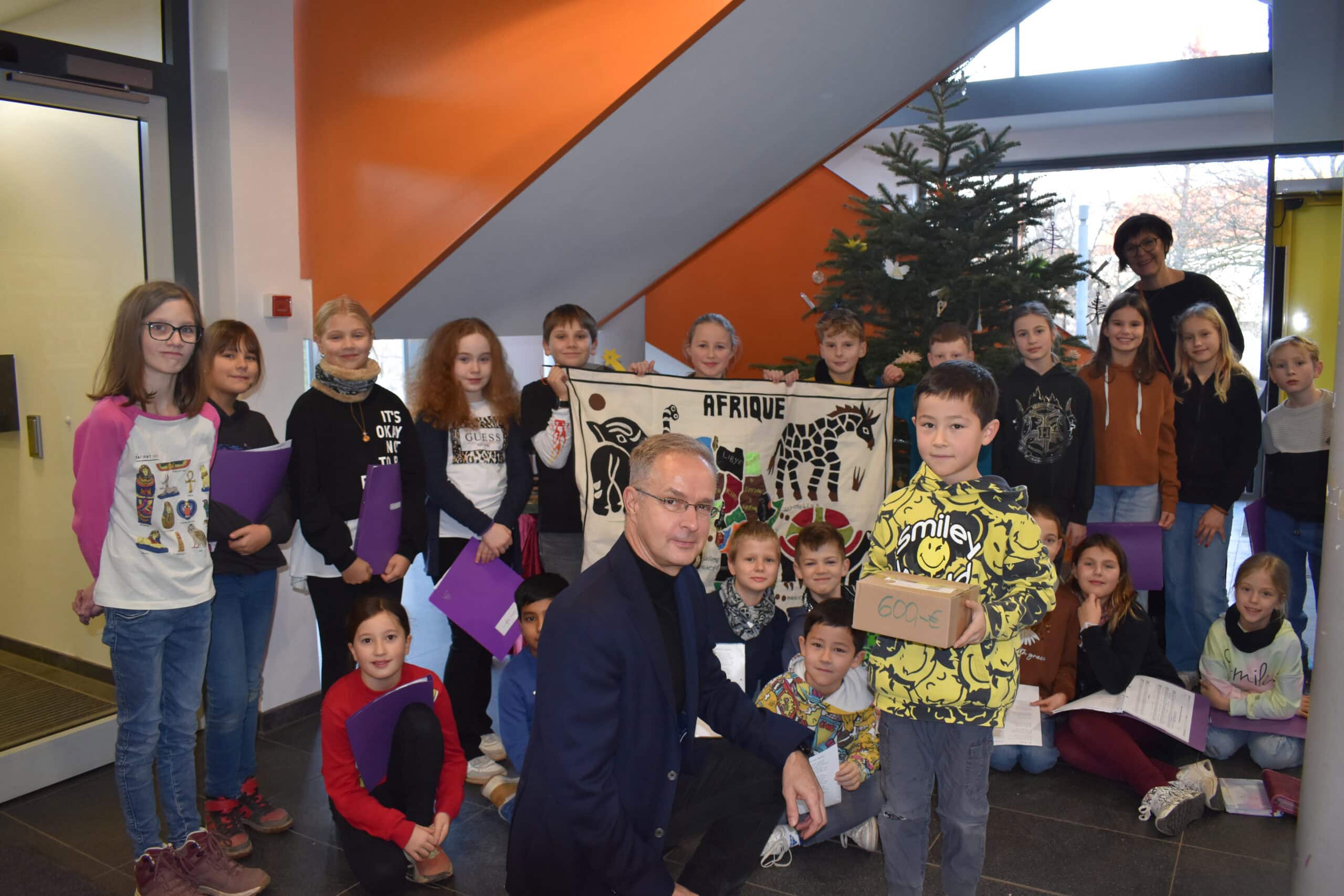 Eine Gruppe von Kindern mit einem Mann, der kniet und eine Kiste hält, neben einem Weihnachtsbaum.