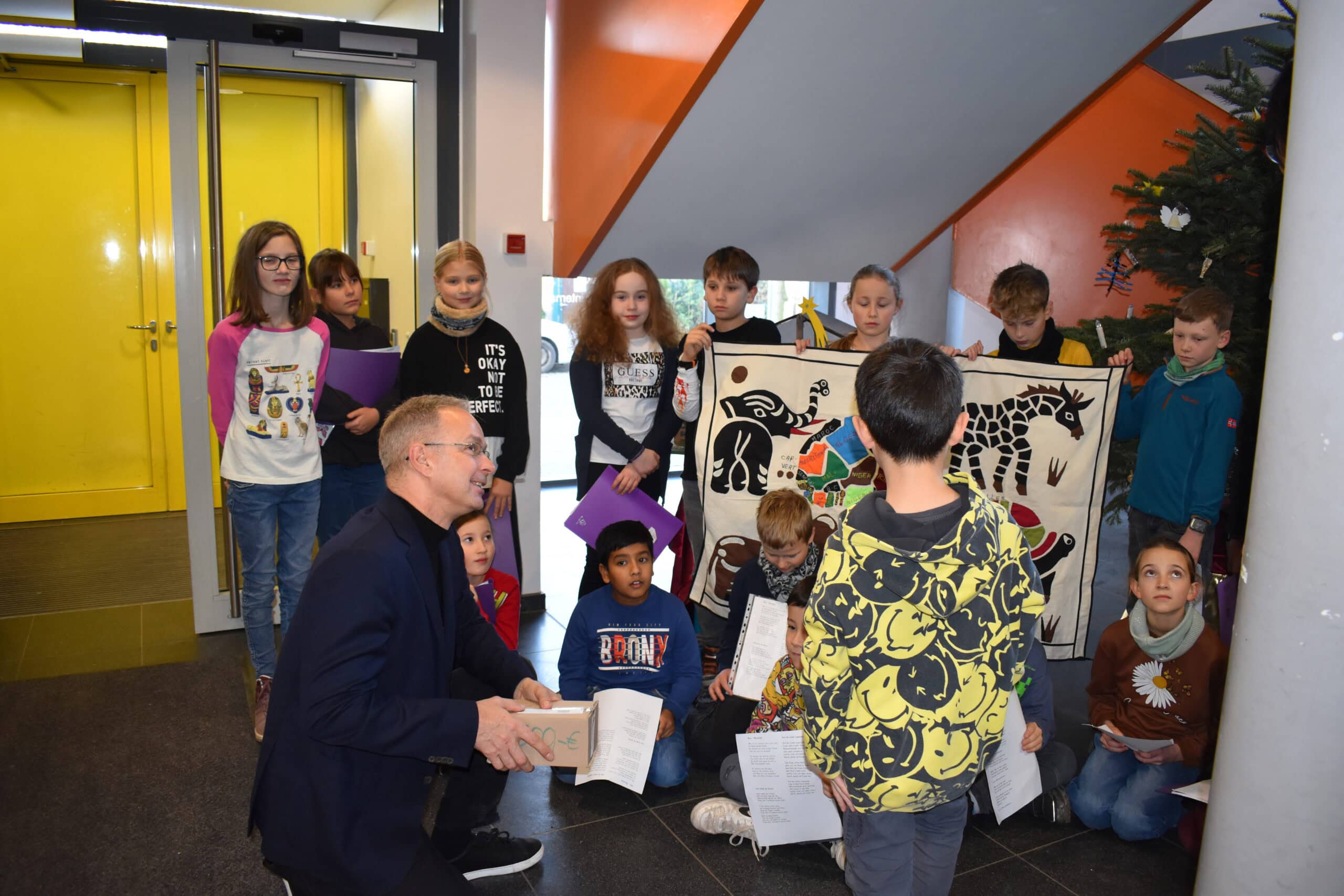 Eine Gruppe Kinder singt fröhlich, ein Mann lächelt ihnen zu, bunte Kleidung, weihnachtlicher Hintergrund.