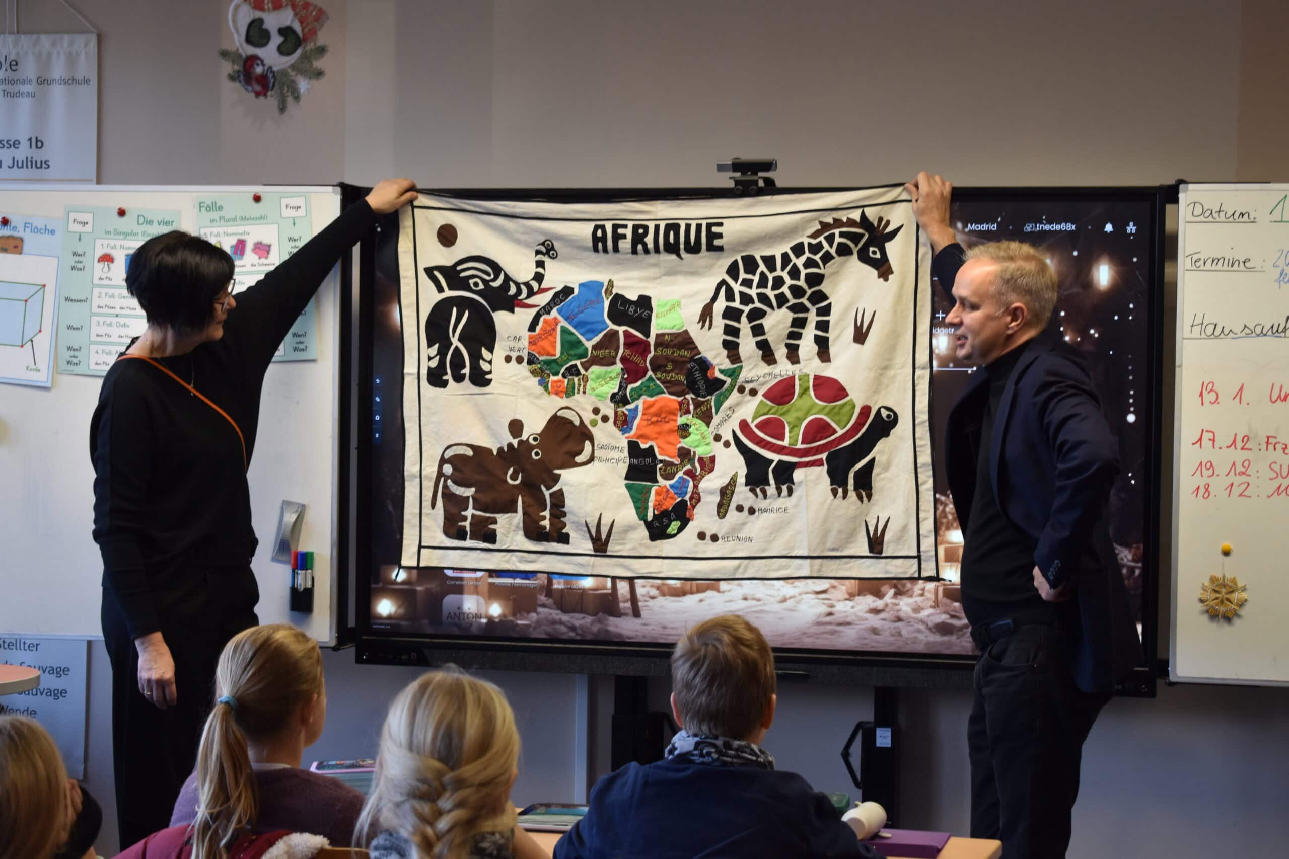 Zwei Erwachsene zeigen Kindern ein buntes Afrika-Poster mit Tiermotiven in einem Klassenzimmer.