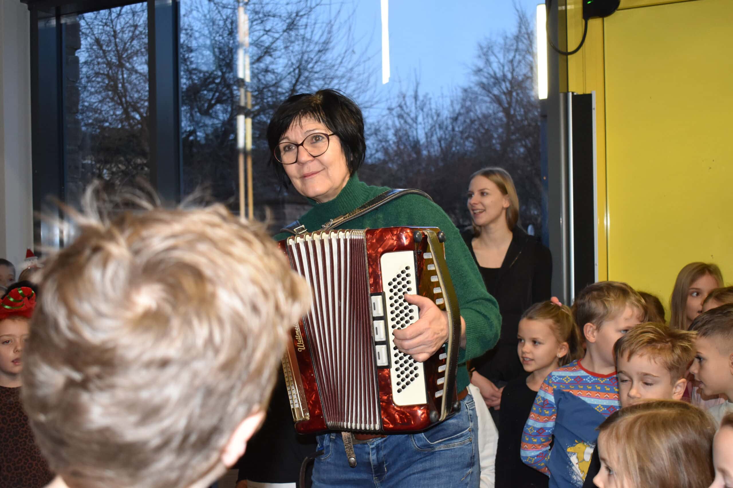 Eine ältere Frau spielt Akkordeon bei einer fröhlichen Kindergruppe; Hintergrund: Glasfenster.