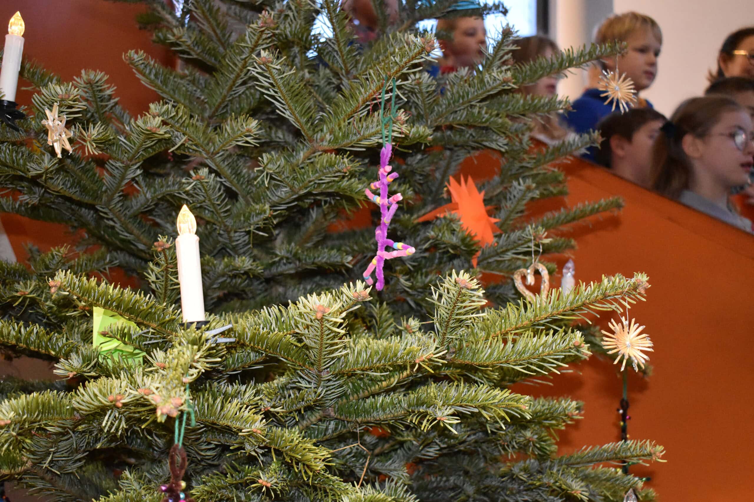 Ein geschmückter Weihnachtsbaum mit Kindern im Hintergrund. Grüne Zweige, bunte Dekorationen.