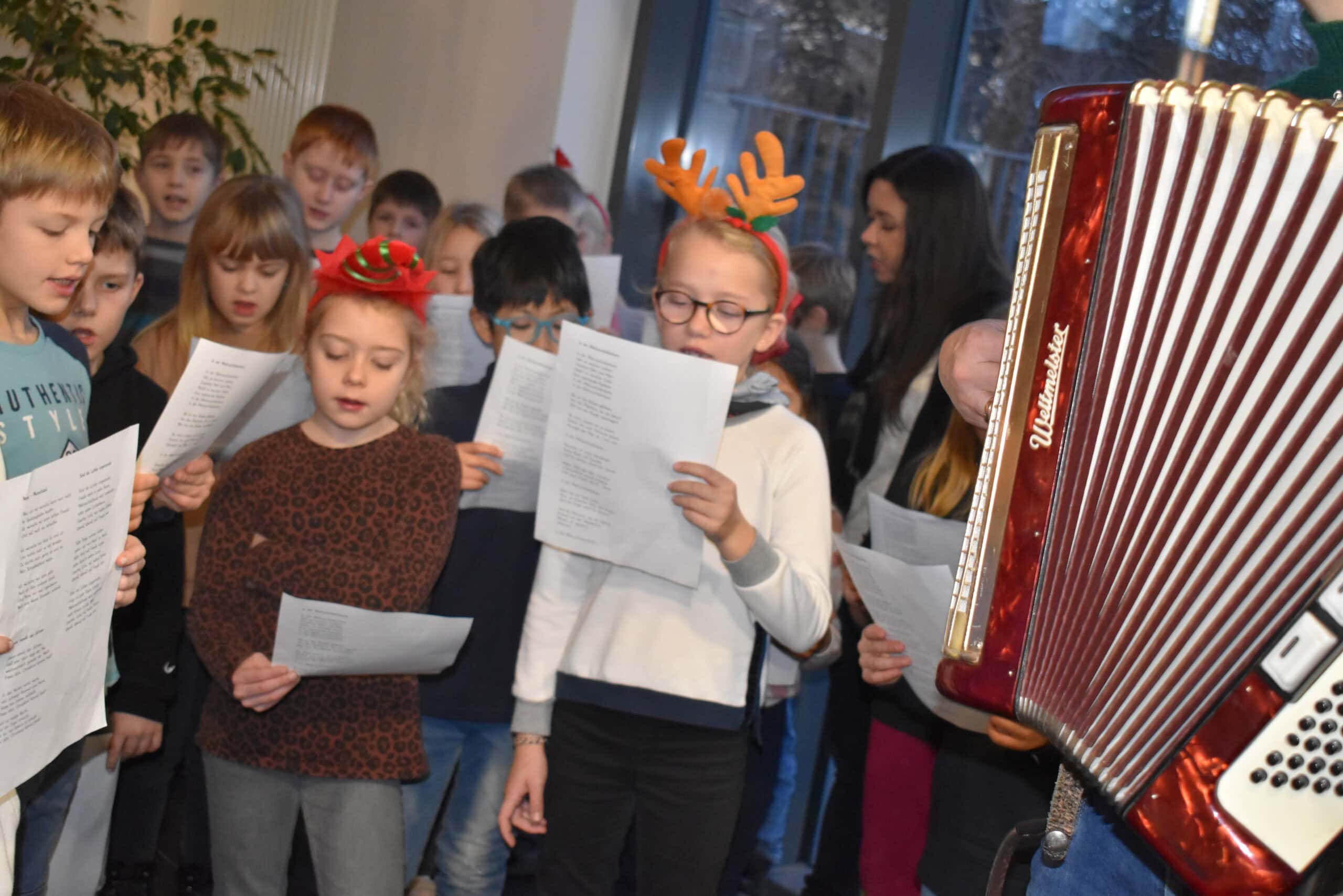 Kinder singen Weihnachtslieder, einige tragen Rentiergeweihe. Akkordeon im Vordergrund.