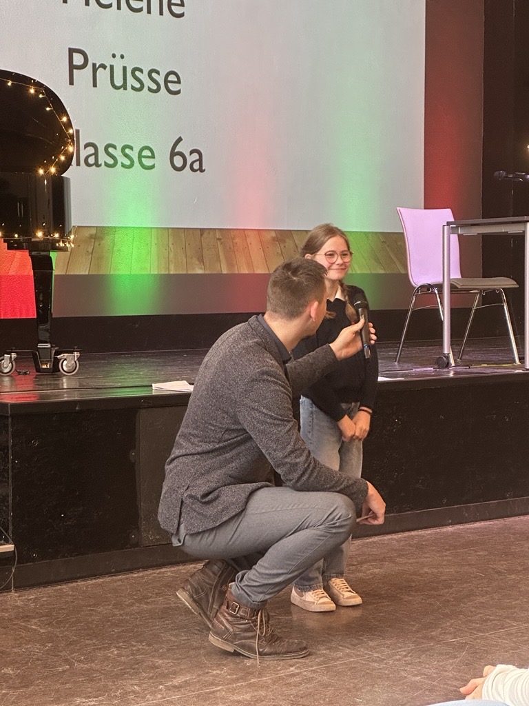 Erwachsener kniet und spricht mit lächelndem Kind auf Bühne; bunte Bühne im Hintergrund.