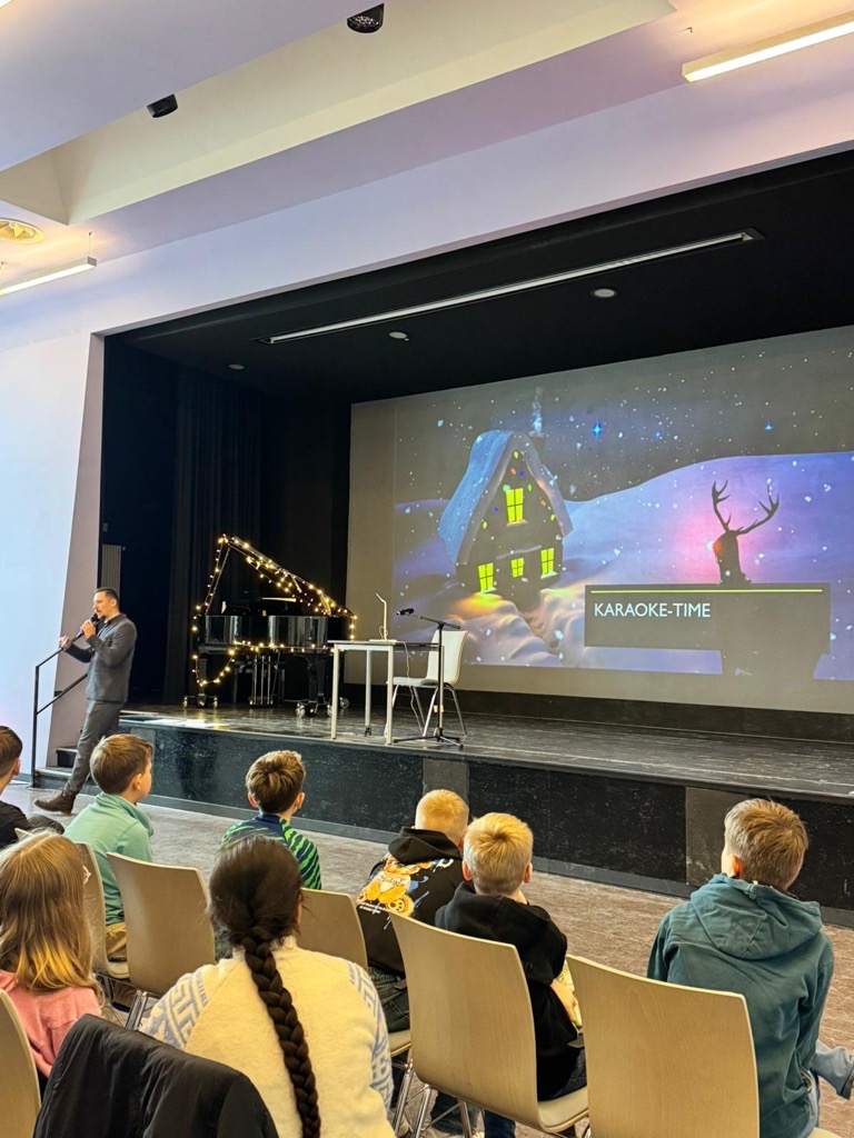 Ein Mann auf Bühne mit Mikrofon, Kinder sitzen davor; Karaoke-Zeit, dunkle Bühne, winterliches Motiv.