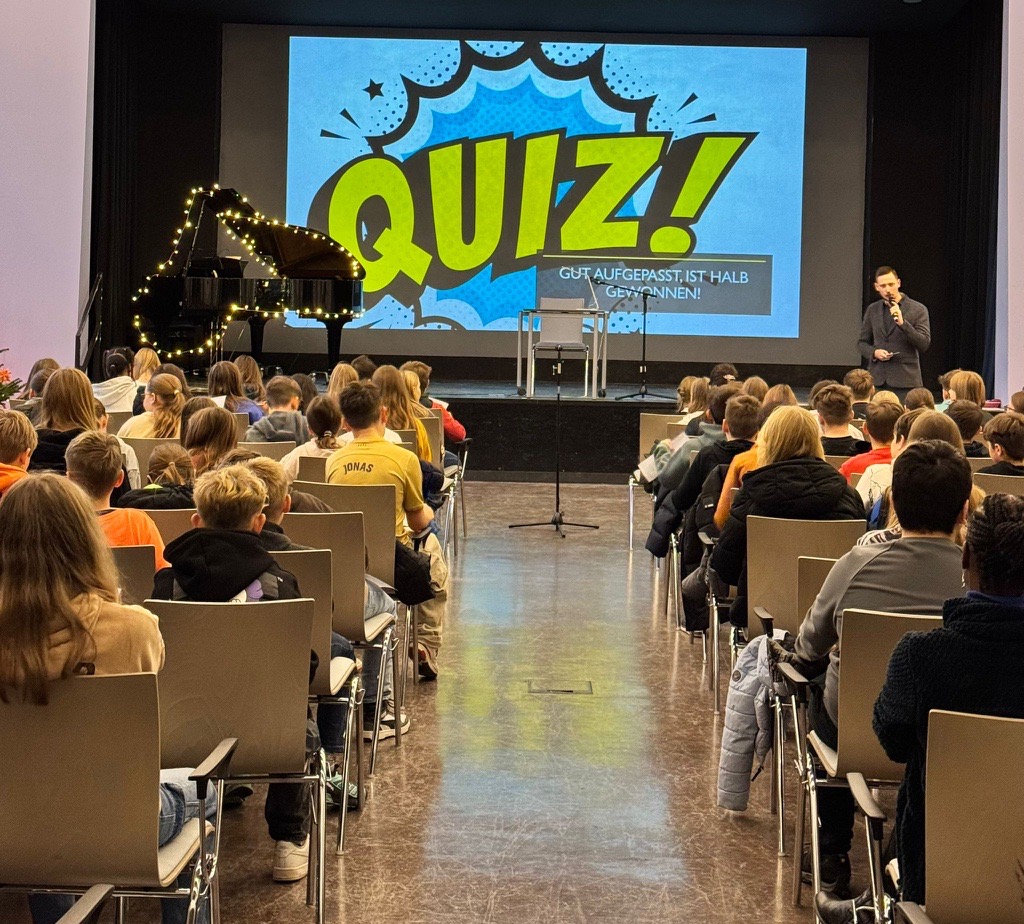 Eine Gruppe Jugendlicher sitzt, während ein Mann neben einer Leinwand mit dem Wort "QUIZ" steht.