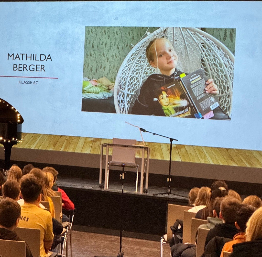 Ein Mädchen im Bild hält ein Buch; lächelnd. Auf Projektor präsentiert. Publikum schaut zu.