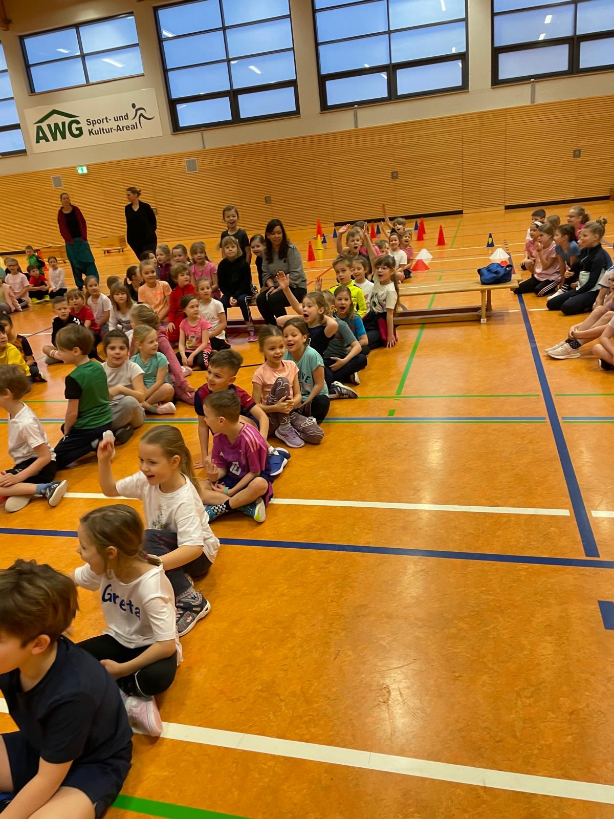 Kinder in Turnhalle, fröhlich, sitzen in Reihen auf dem Boden, Sportkleidung, bunte Kegel hinten.