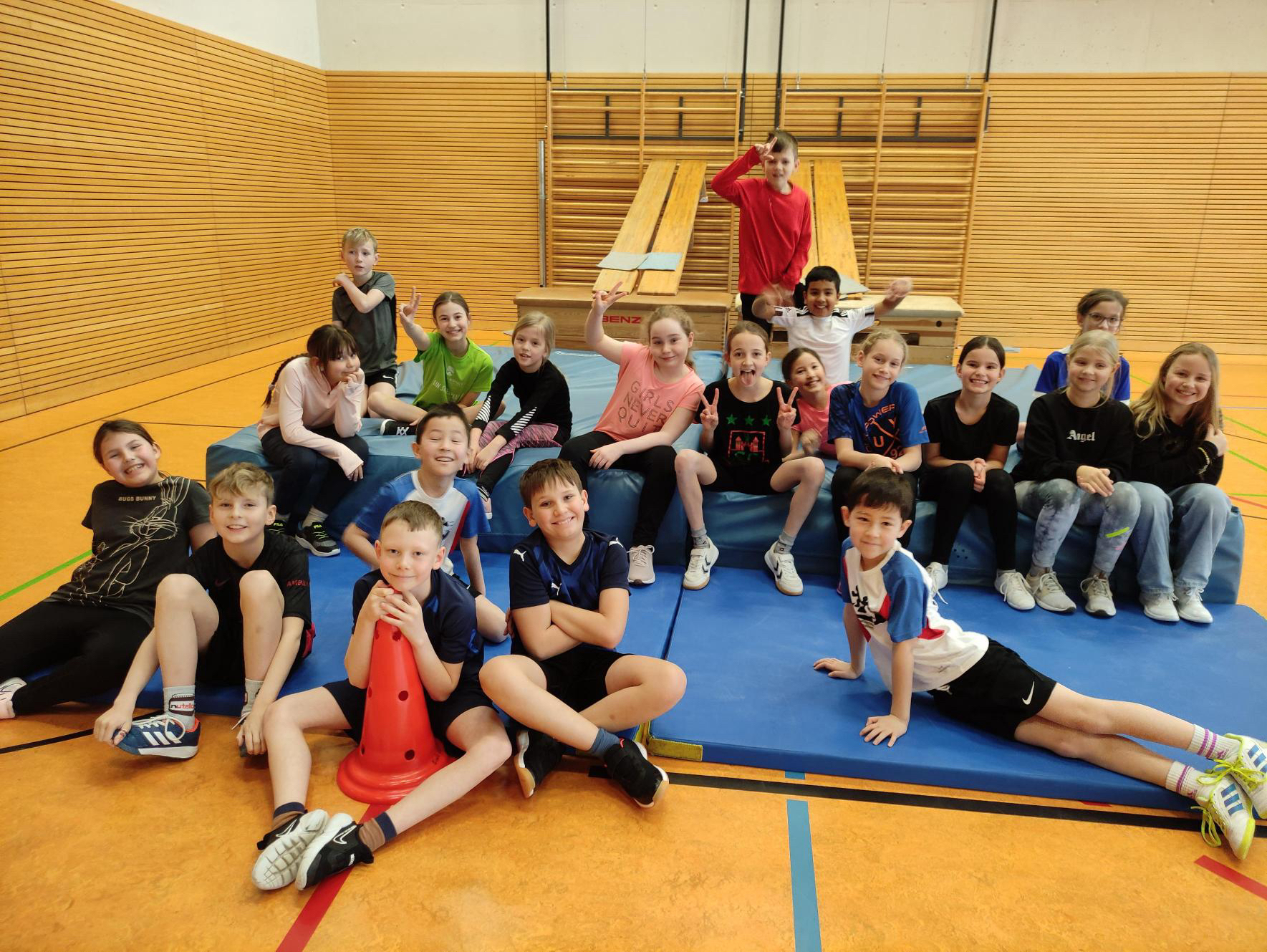 Gruppe fröhlicher Kinder in Sporthalle, sitzt auf Matten, bunte Sportkleidung, hölzerne Wand.