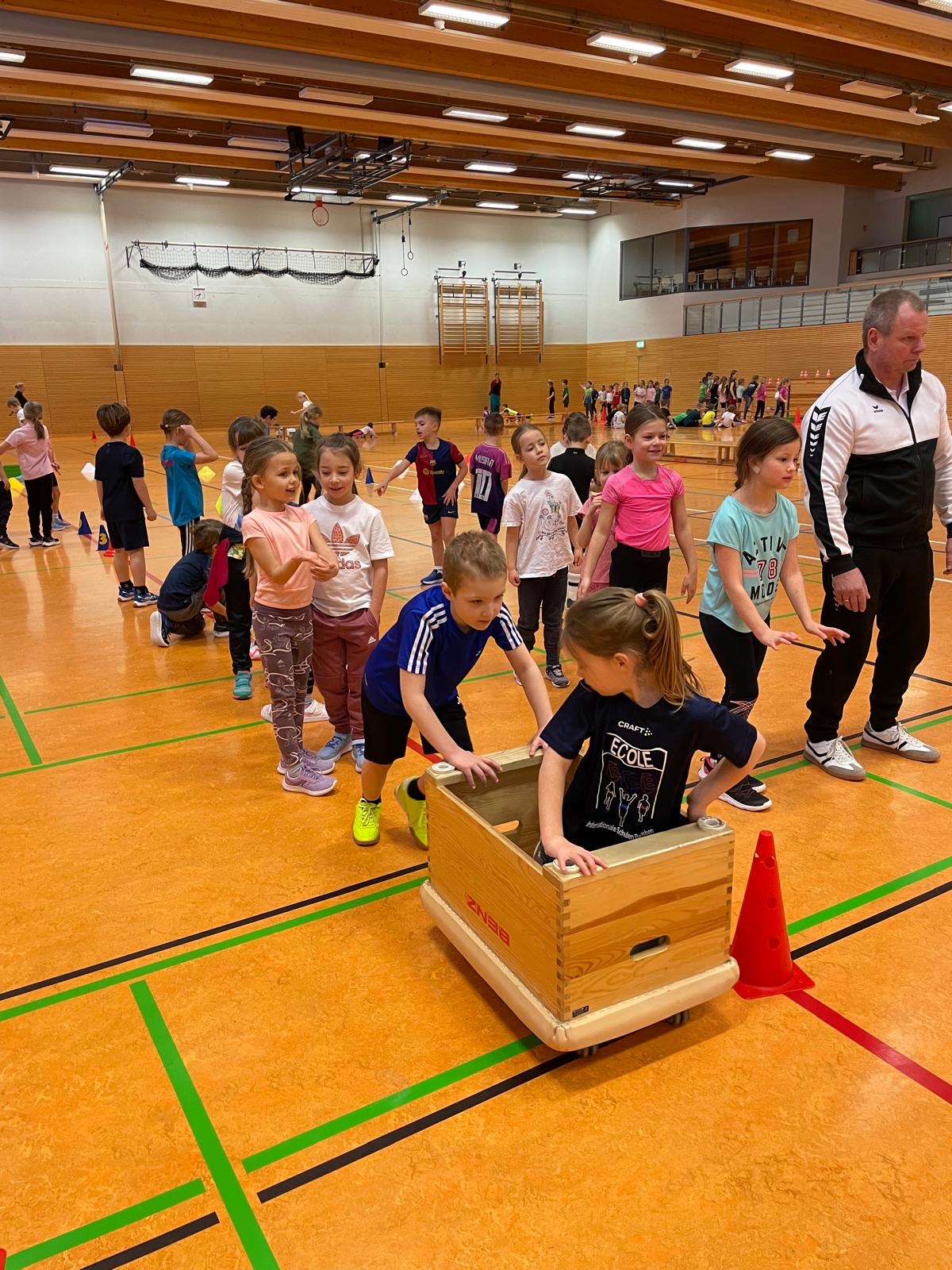 Kinder spielen in Turnhalle, lächeln, tragen bunte Sportkleidung, eine Kiste wird geschoben.