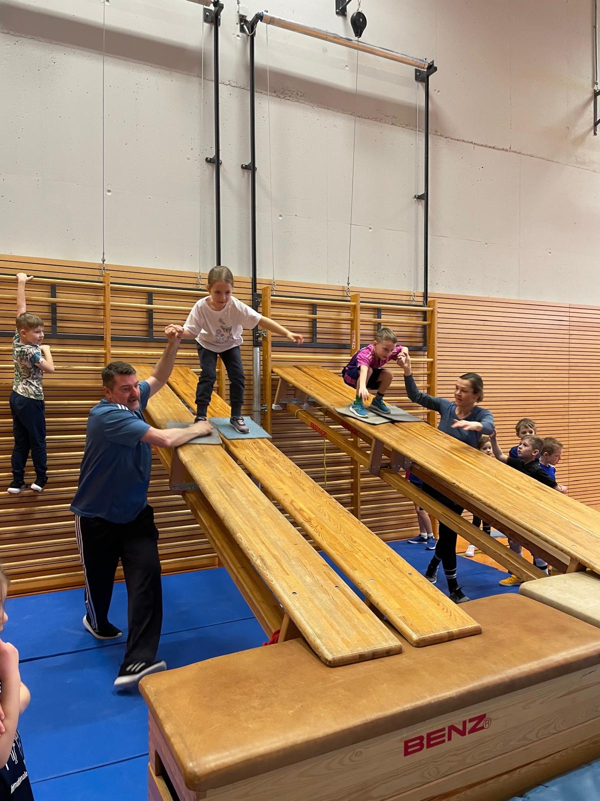 Eine Gruppe von Kindern balanciert auf Turnbänken in einer Sporthalle. Ein Mann hilft einem Kind.