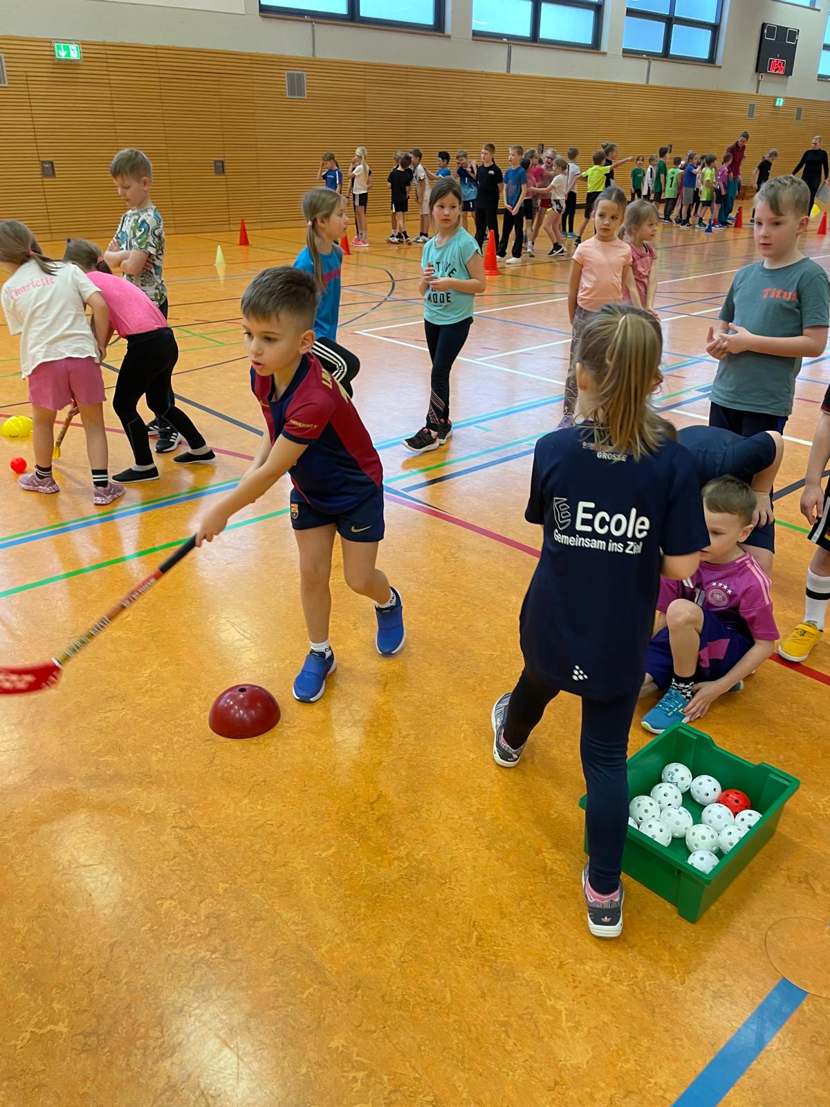 Kinder beim Hallensport, fröhlich, tragen farbige Trikots, spielen Kurzhockey, helle Halle.