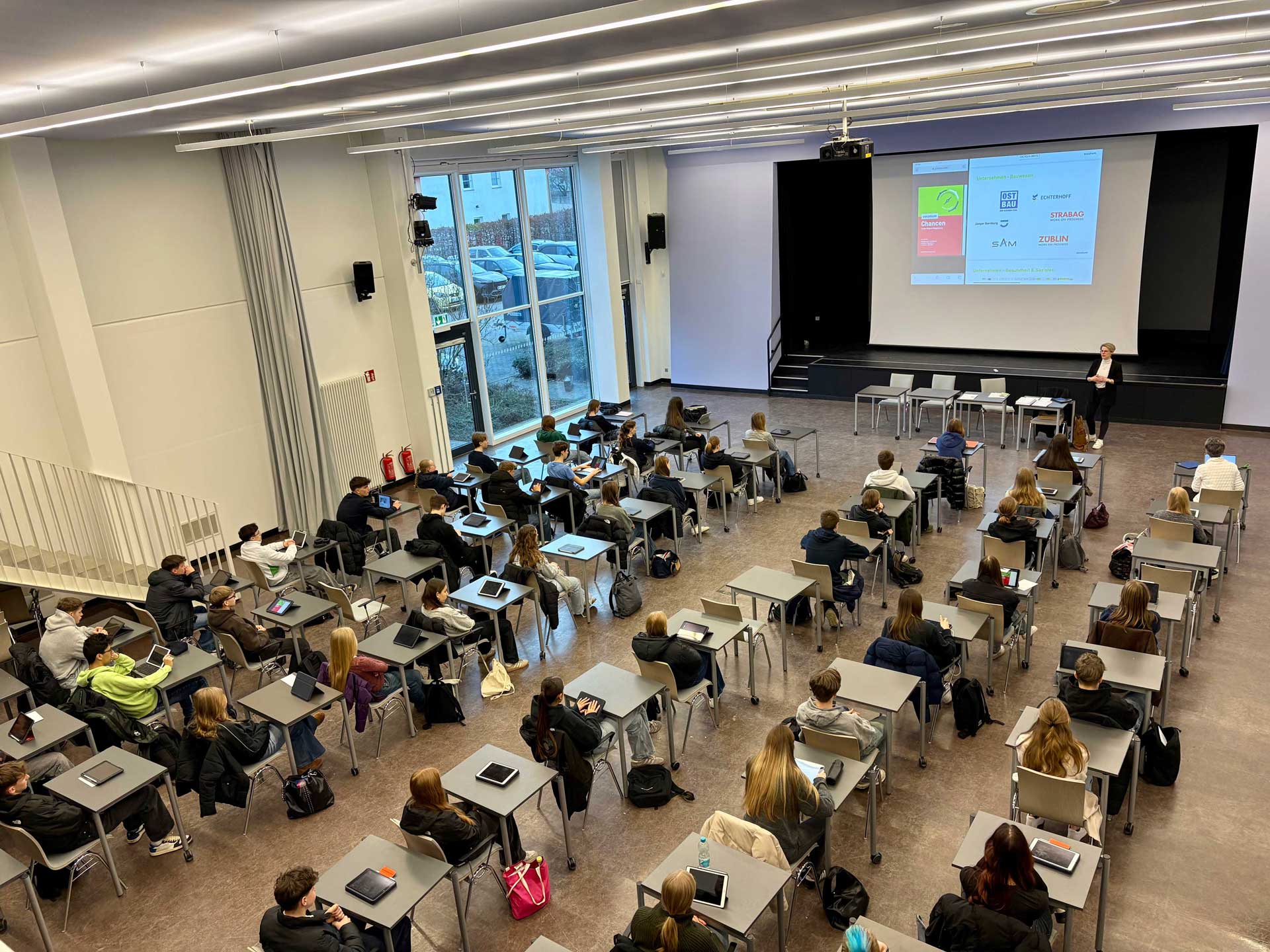 Ein Klassenzimmer mit sitzenden Personen, die einem Vortrag vor einer Leinwand folgen.