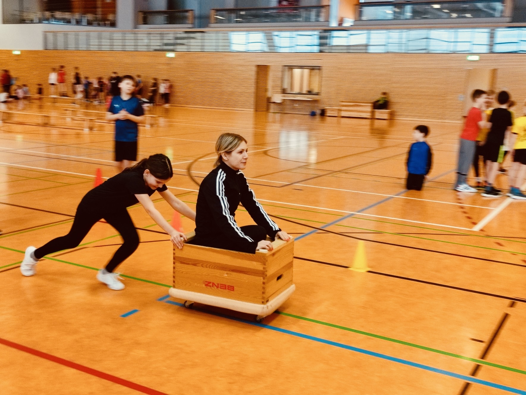 Eine Frau wird lachend in einer Turnhalle auf einem Holzkasten geschoben, Jugendliche im Hintergrund.