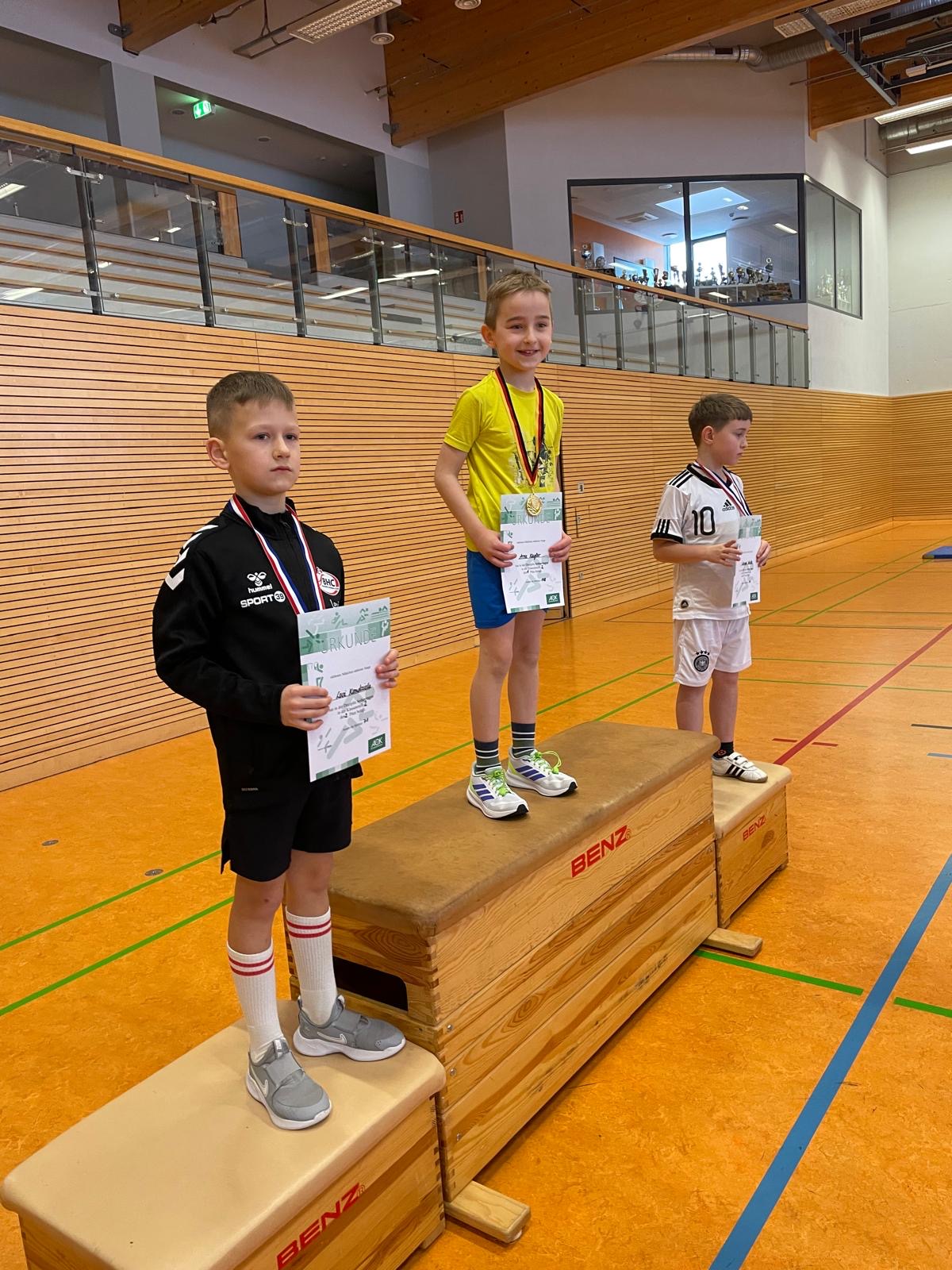 Drei Kinder, fröhlich, auf Siegerpodest mit Medaillen in Sporthalle, Holzverkleidung, leuchtende Farben.