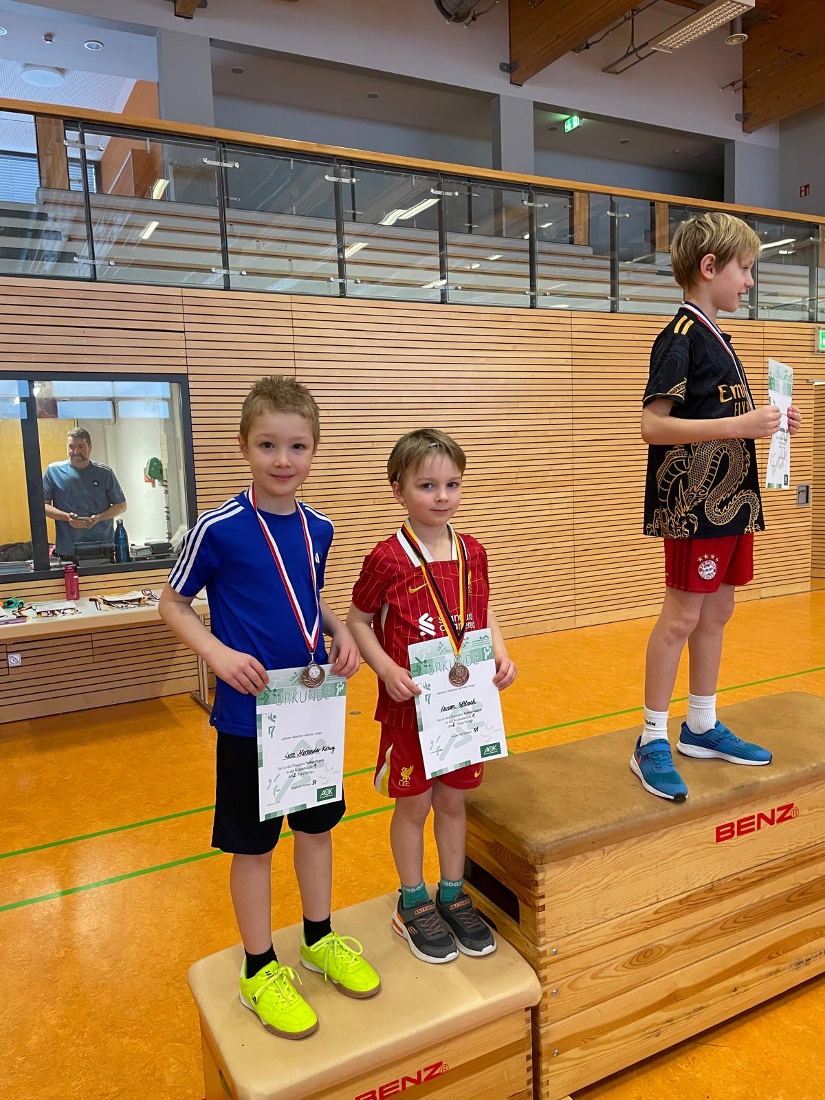 Zwei Kinder mit Medaillen und Urkunden auf Podest, lächelnd in Sportkleidung, in Turnhalle.