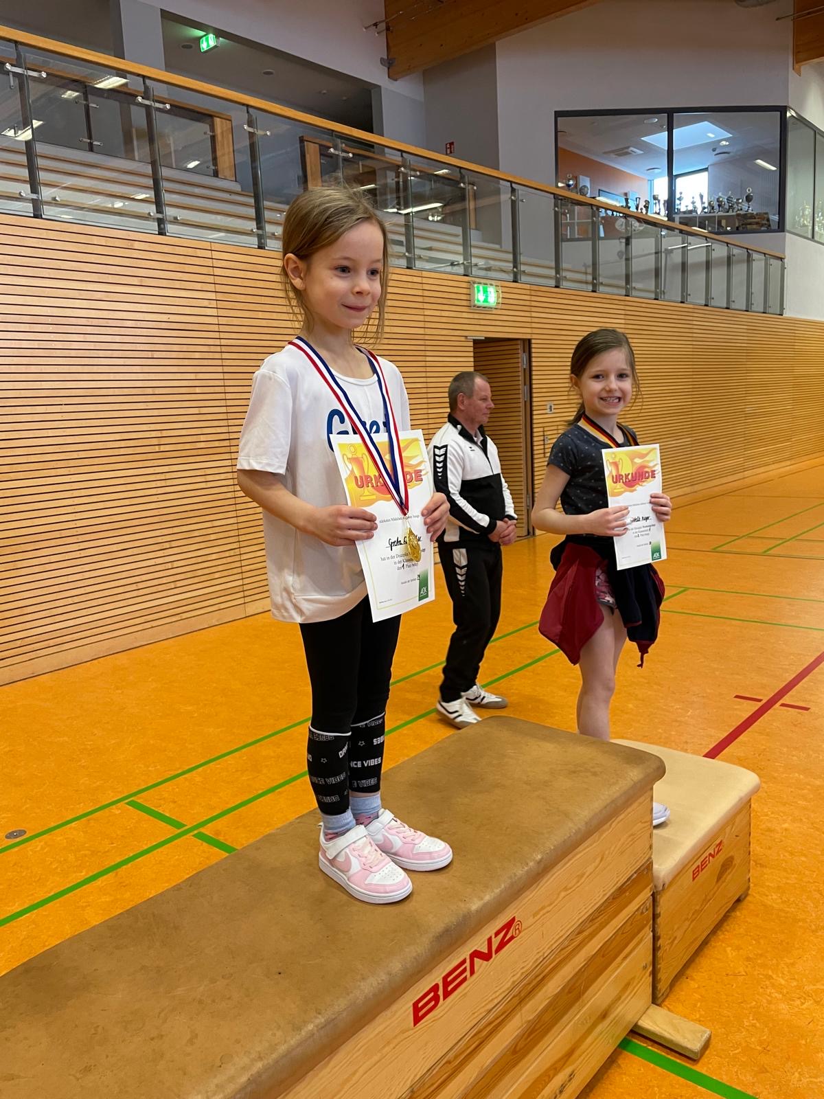 Zwei Mädchen auf einem Siegerpodest, die Medaillen und Urkunden halten, lächelnd, in Sporthalle.