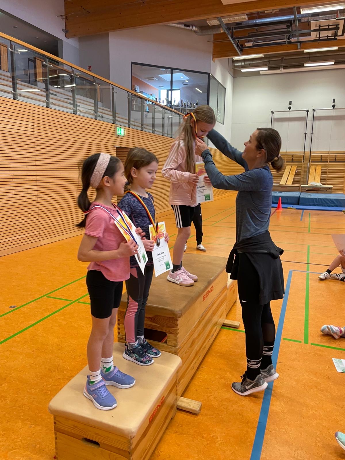 Eine erwachsene Frau verleiht drei Mädchen mit Medaillen auf Holzpodesten in einer Sporthalle.