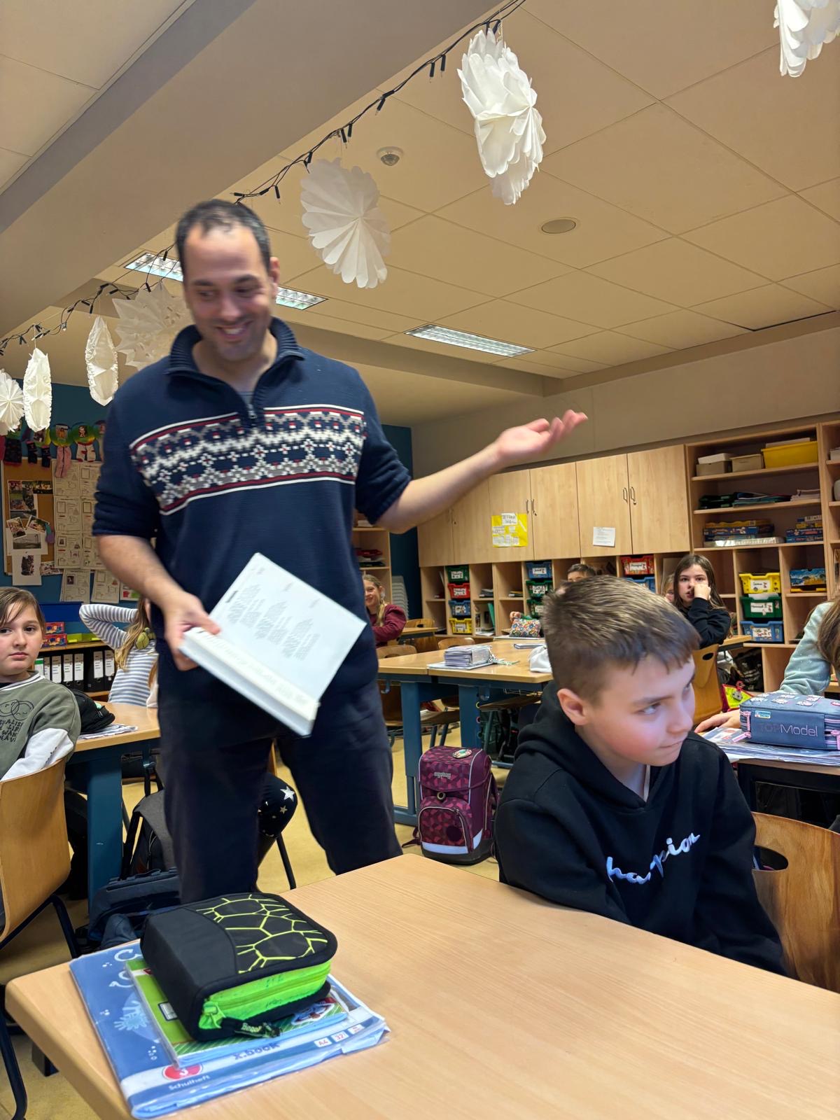 Ein lächelnder Mann hält ein Buch in einem Klassenzimmer; Kinder sind an Tischen.
