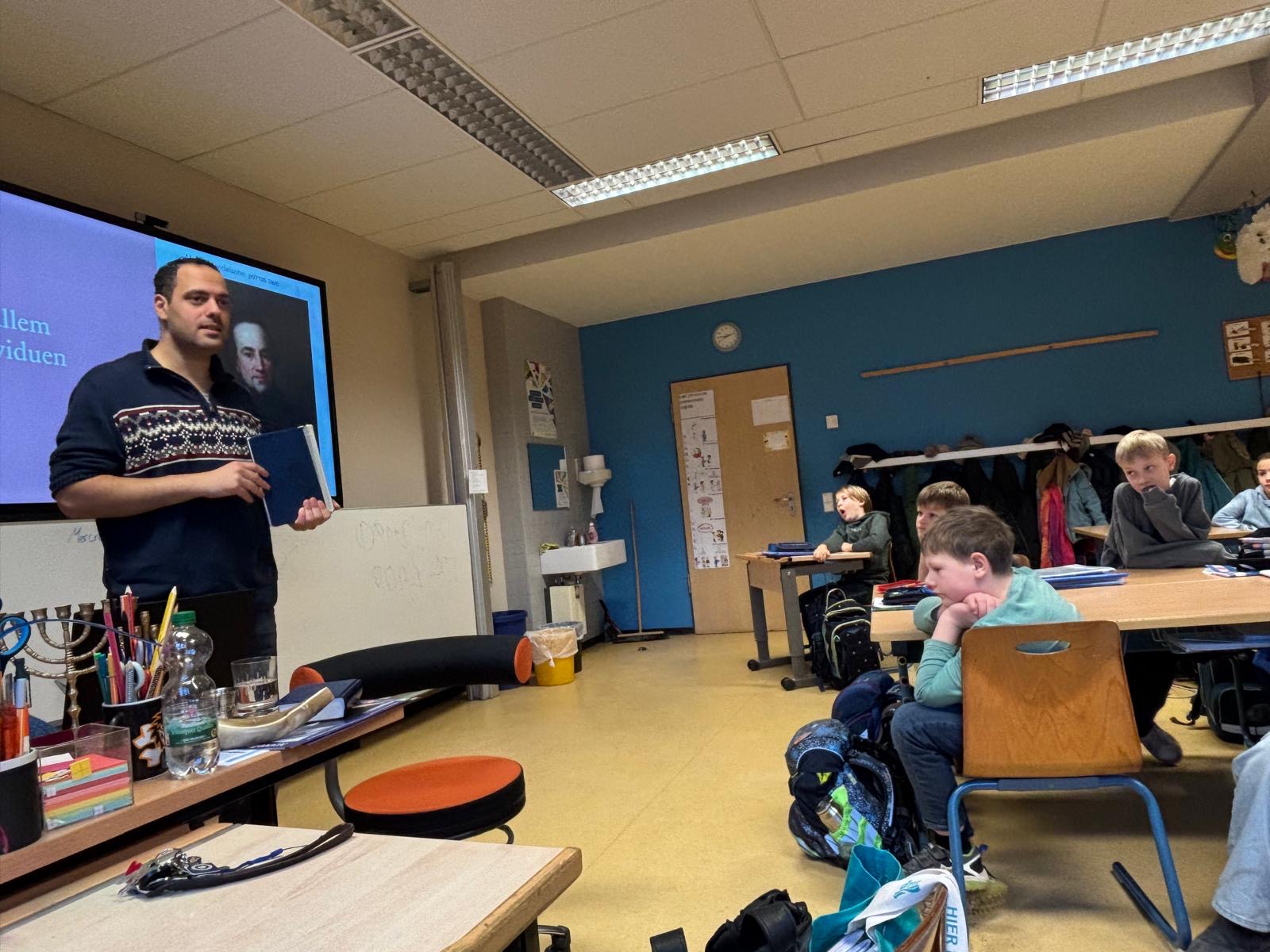 Ein Lehrer erklärt vor der Klasse; Schüler hören zu; bunte Wände, Holzschreibtische, konzentration.