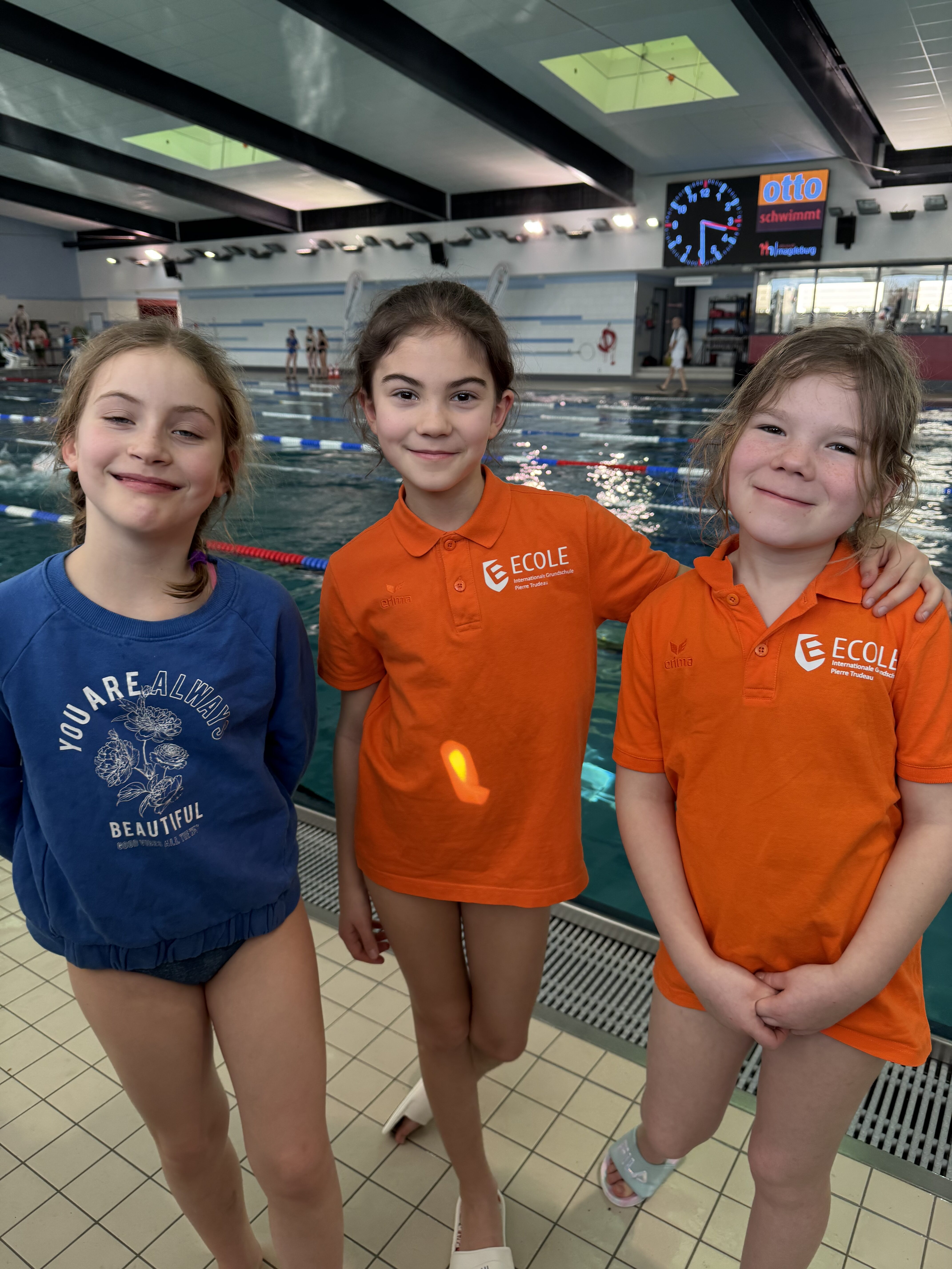 Drei lächelnde Mädchen am Pool in blauer und orangener Kleidung. Sportlicher Hintergrund.