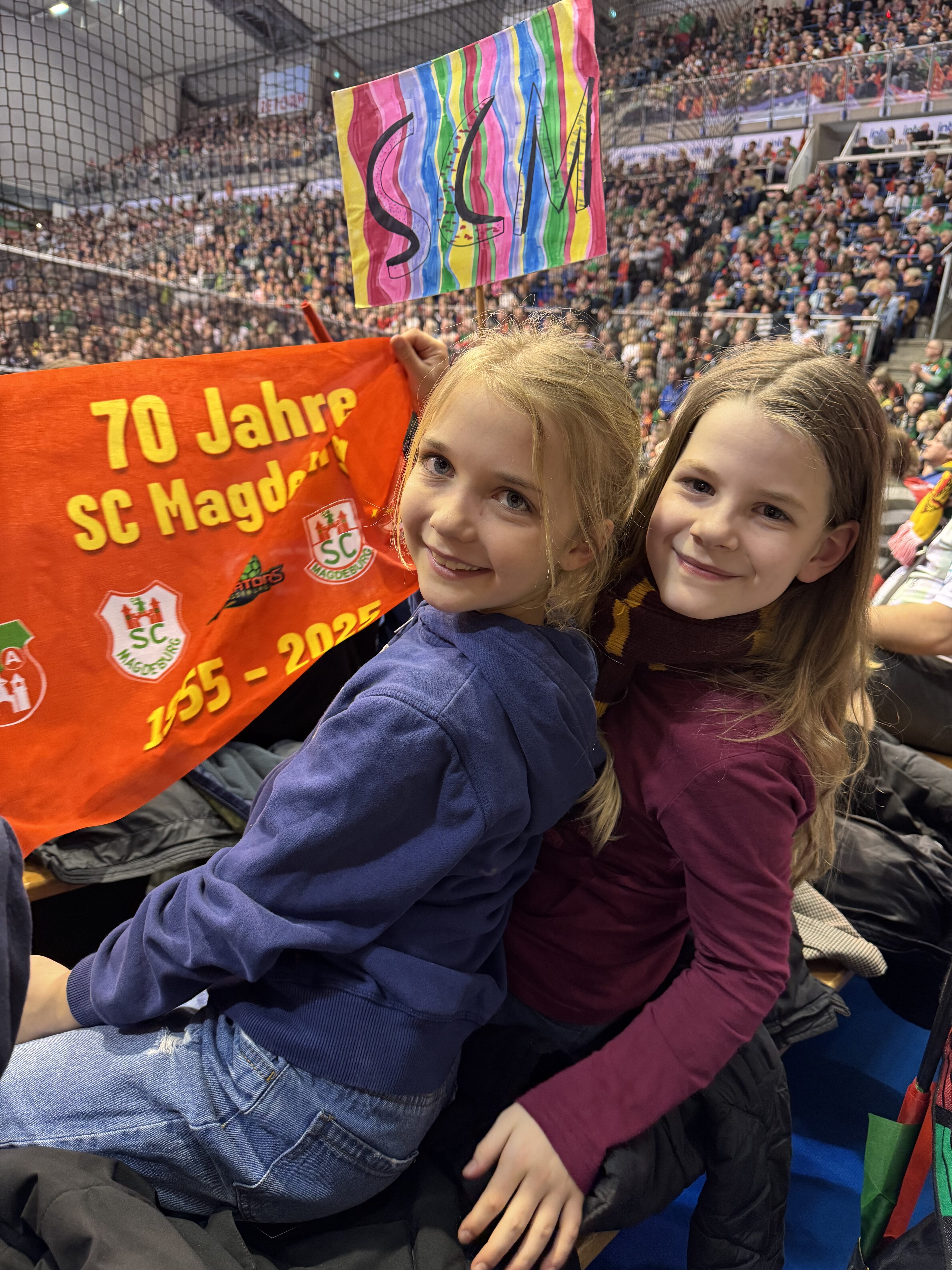 Zwei Mädchen lächeln, halten ein buntes Schild und ein rotes Jubiläumsbanner in einer vollen Arena.