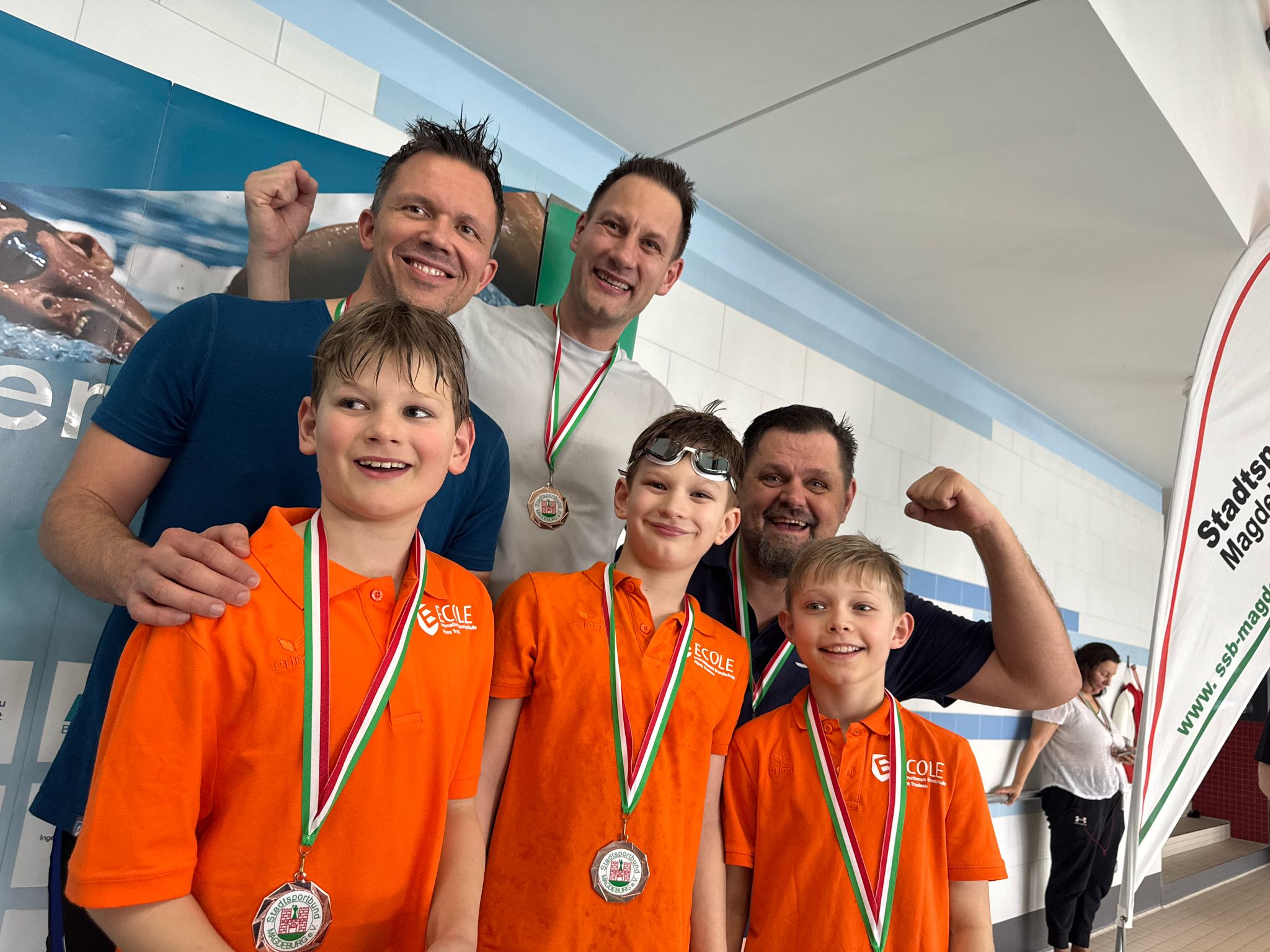 Drei Jungen in orangefarbenen Hemden mit Medaillen, zwei Erwachsene jubeln im Hintergrund.