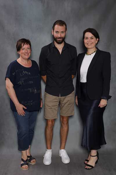 Drei Personen lächeln vor grauem Hintergrund; Mann in Shorts, Frauen formal gekleidet.