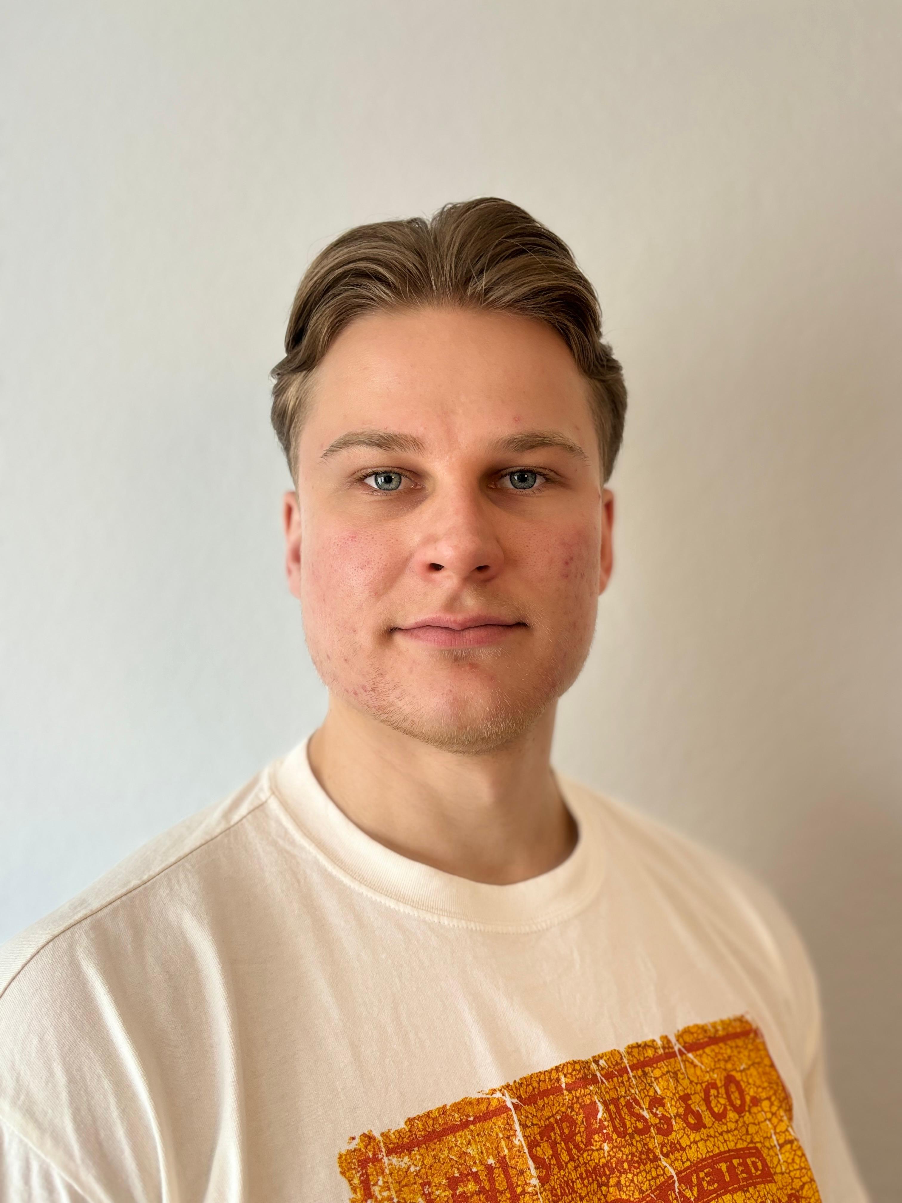 Eine lächelnde Person mit kurzem braunem Haar, weißem Shirt mit rotem Muster, schaut geradeaus.
