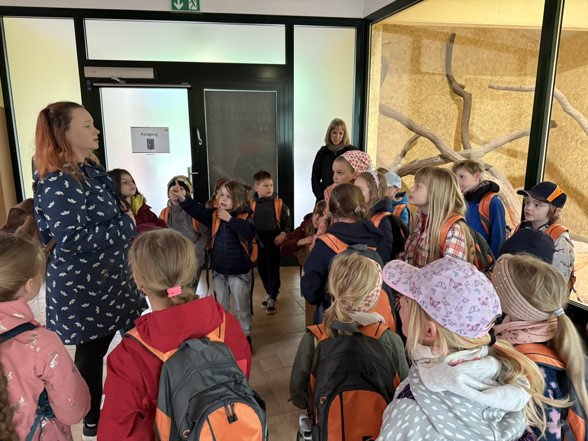 Eine Frau erklärt einer Gruppe von Kindern in Jacken und Rucksäcken; sie hören aufmerksam zu.