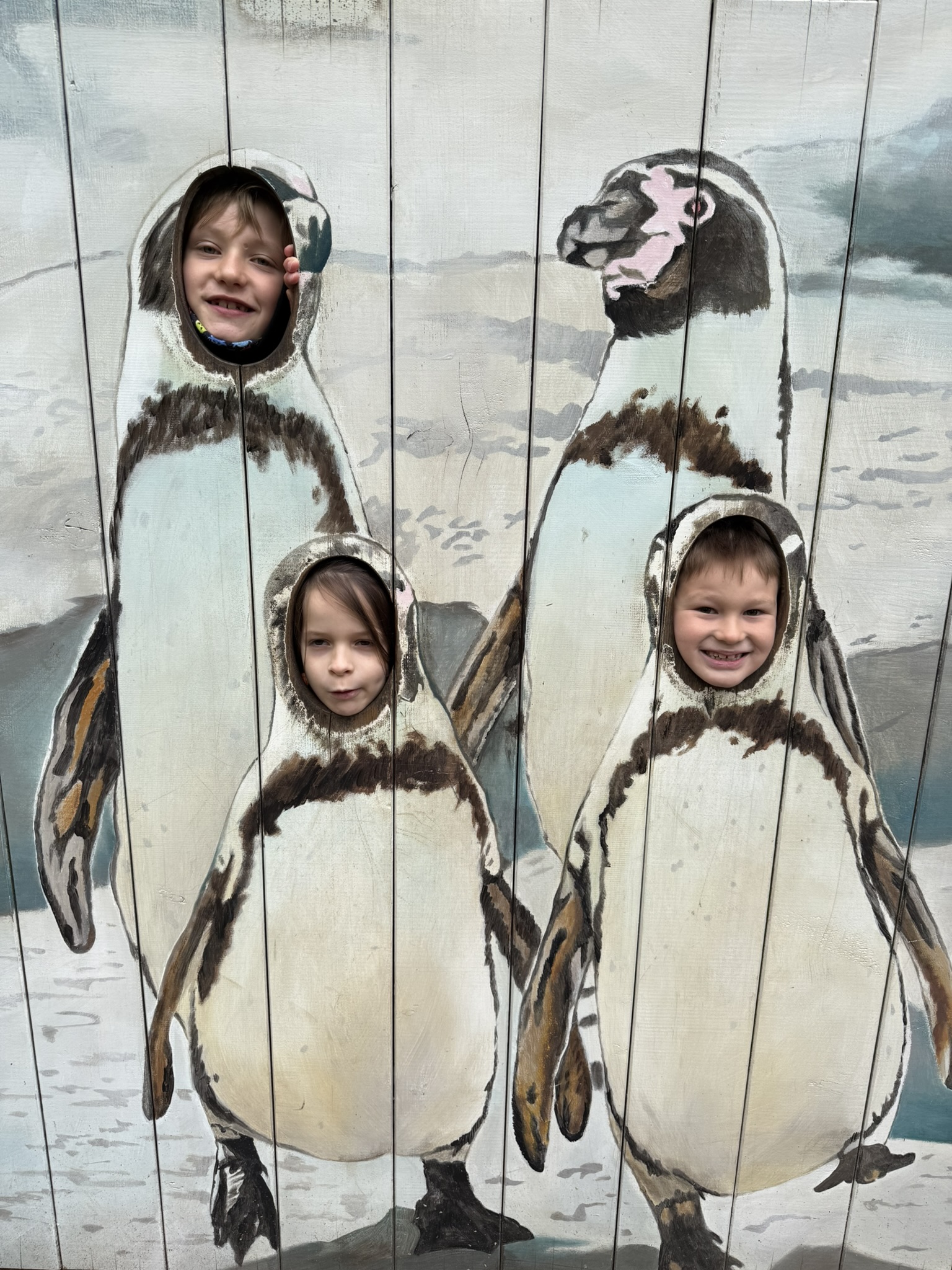 Drei lachende Kinder stecken ihre Köpfe durch Pinguinausschnitte in einer bemalten Holzwand.