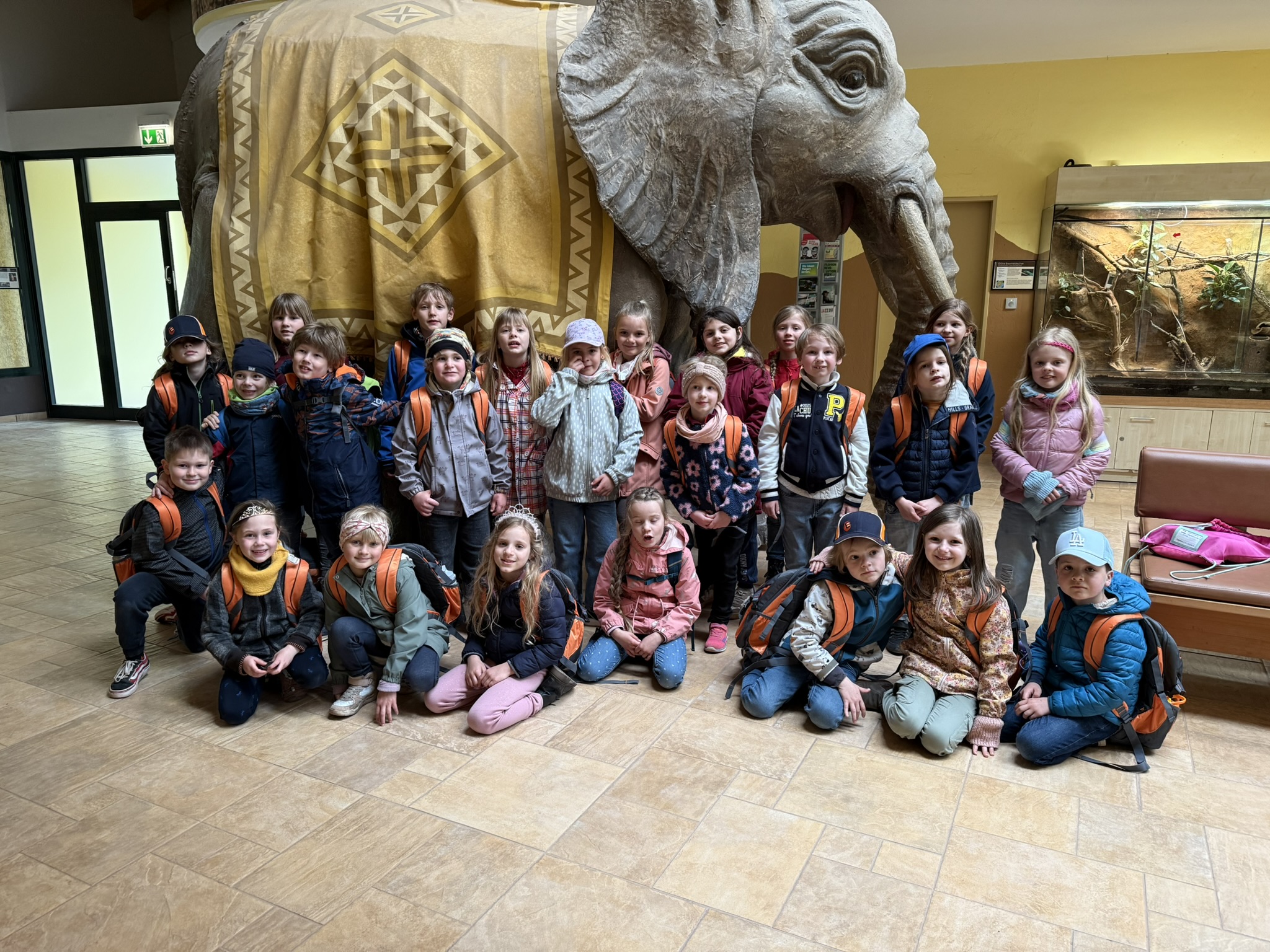 Gruppe von Kindern vor einer großen Elefantenstatue im Gebäude, viele lächeln, bunte Kleidung.