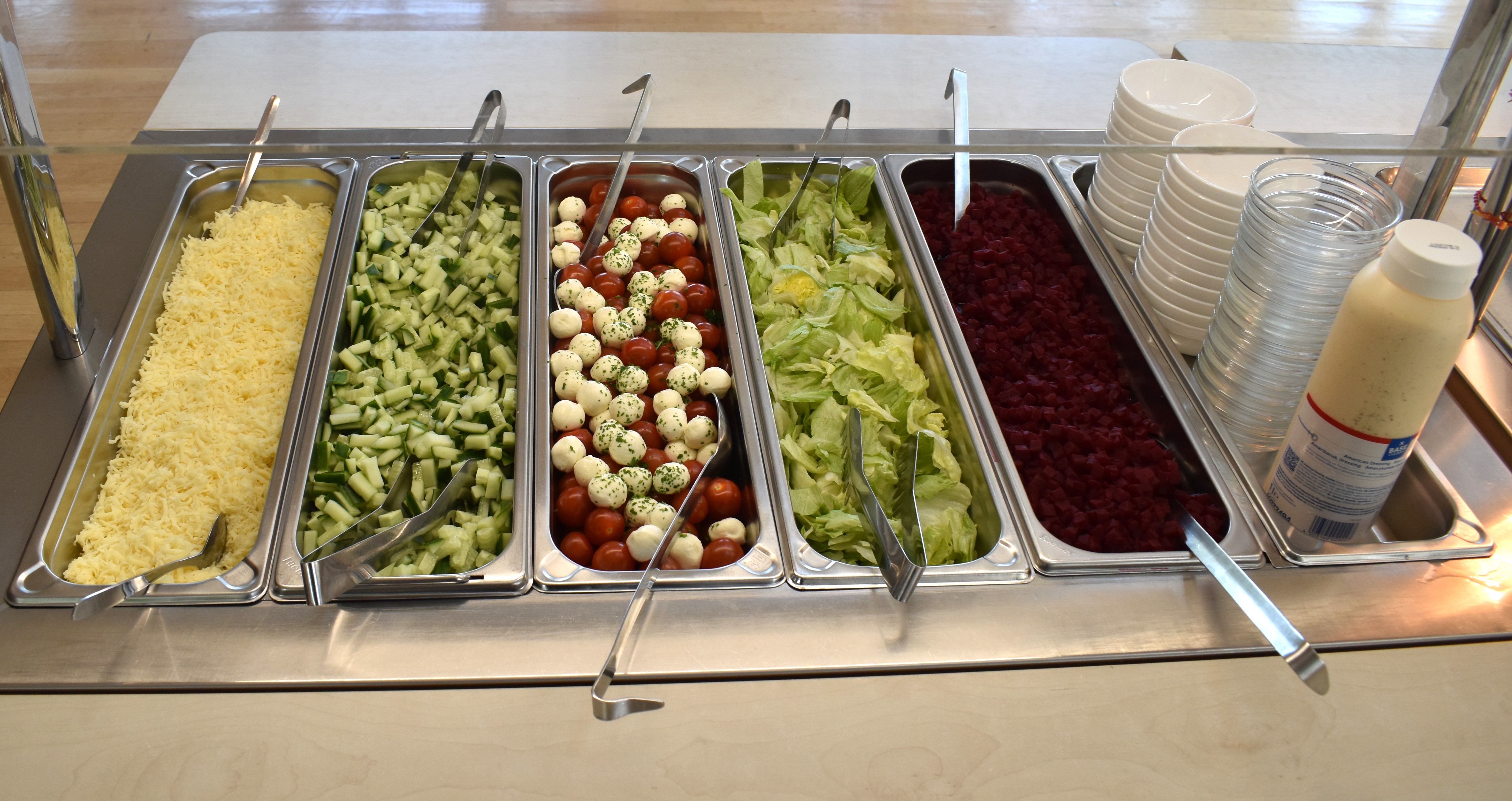 Salatbar mit Käse, Gurken, Tomaten, Moosarella, Salat, Rote Bete, Teller, Becher und Dressingflasche.