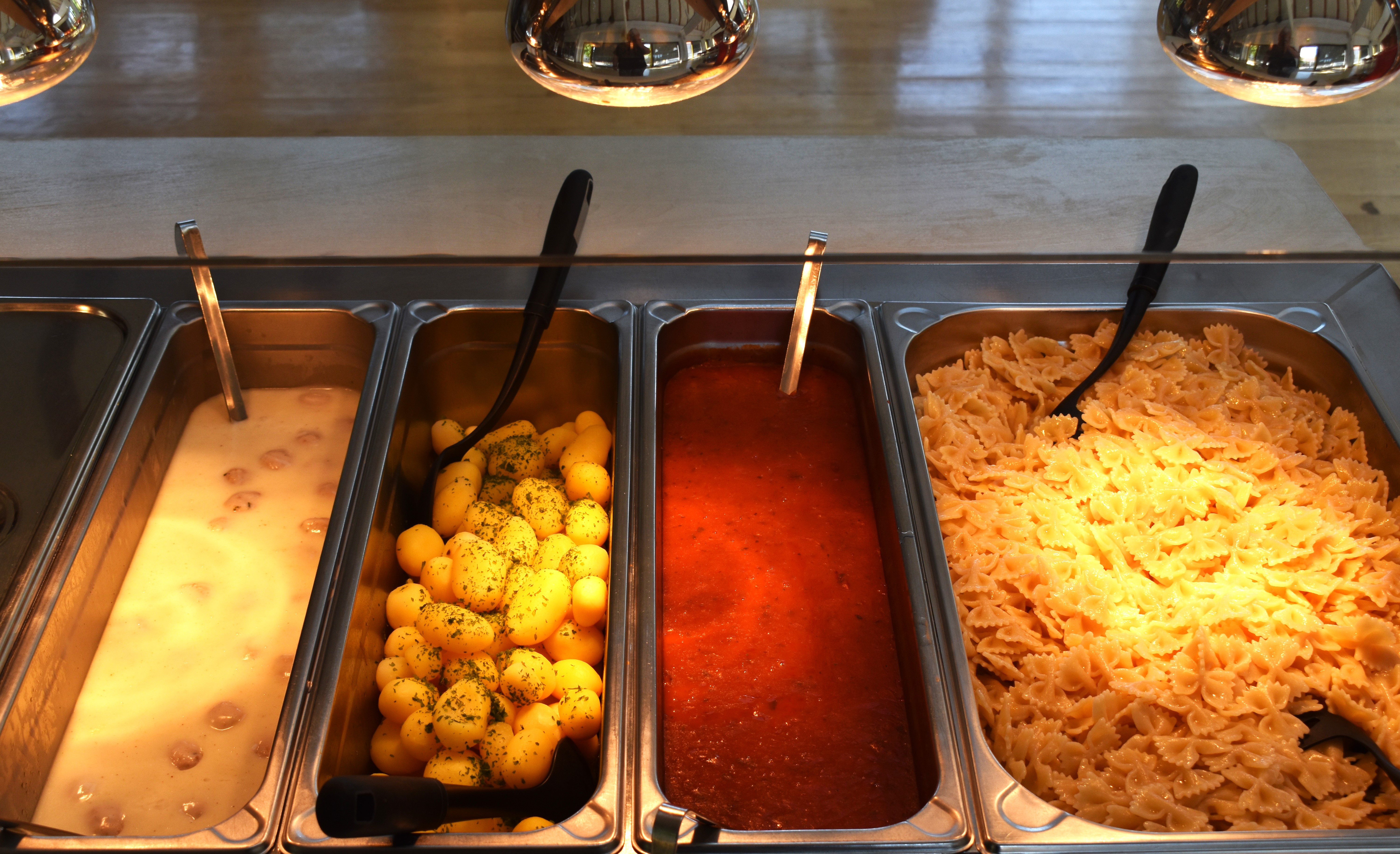 Vier Buffetbehälter mit Soße, Kartoffeln, Tomatensauce und Nudeln unter metallischen Wärmelampen.