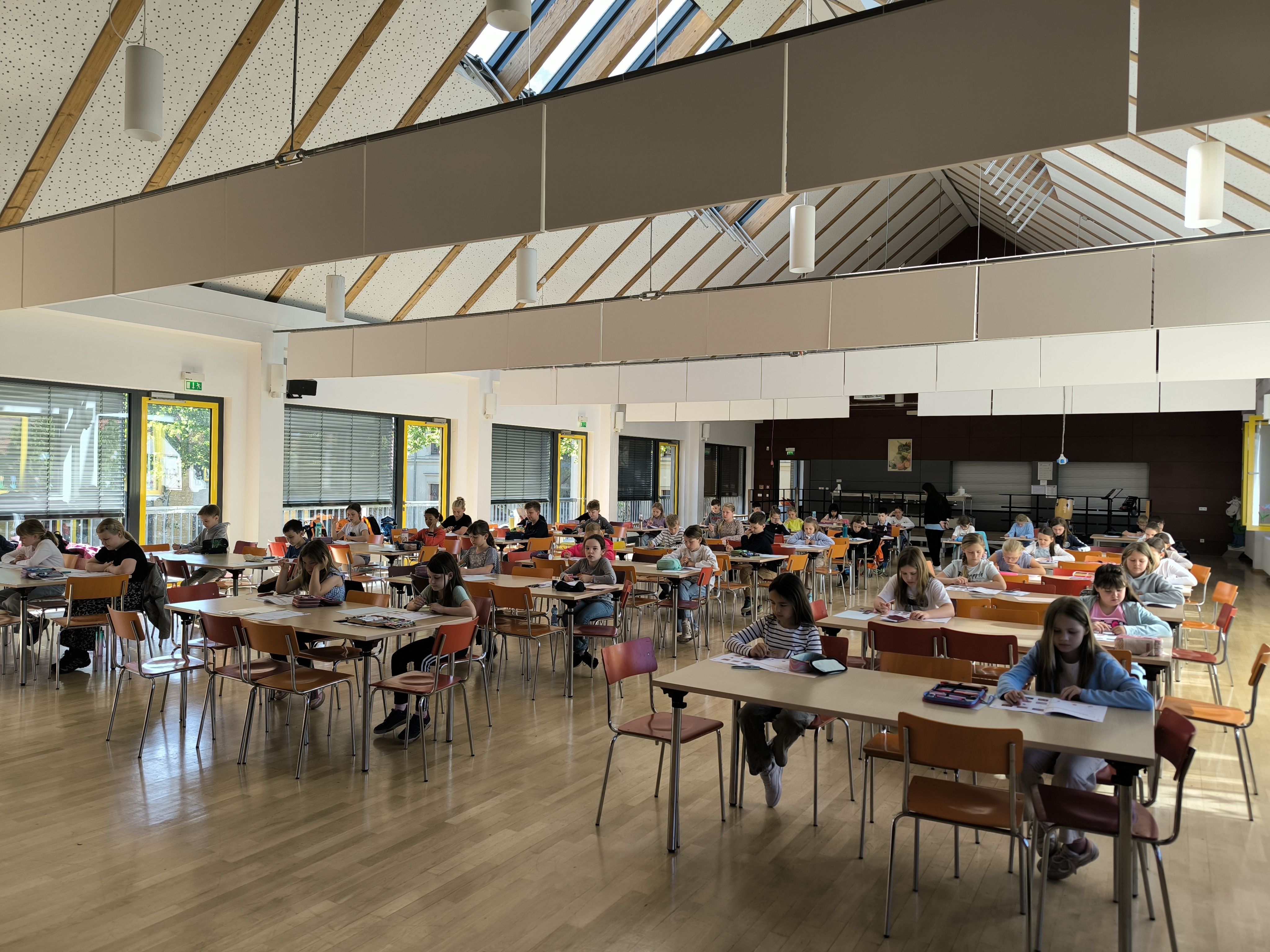 Ein heller, großer Saal mit Schülergruppen an Tischen, die konzentriert arbeiten, Holzboden, Dachfenster.