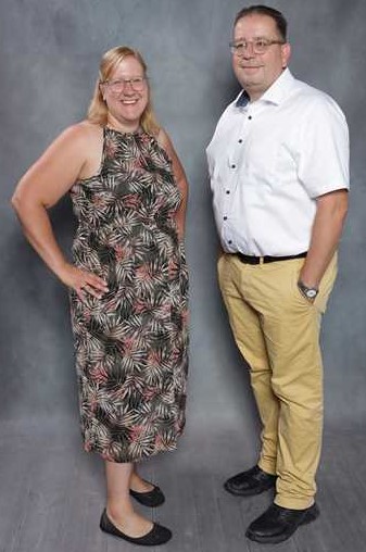 Ein lächelnder Mann in weißem Hemd und eine Frau in gemustertem Kleid posieren vor grauem Hintergrund.