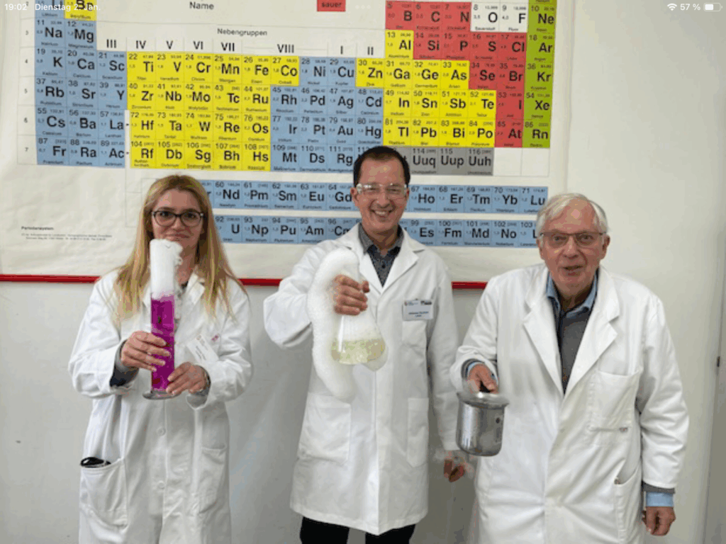 Drei Personen in Laborkitteln mit chemischen Reaktionen vor Periodensystem.