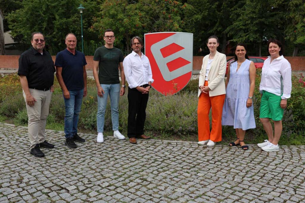 Eine Gruppe von sieben Personen steht lächelnd auf einem Kopfsteinpflaster vor einem Schild mit rotem und grauem Logo, umgeben von Bäumen und Pflanzen.