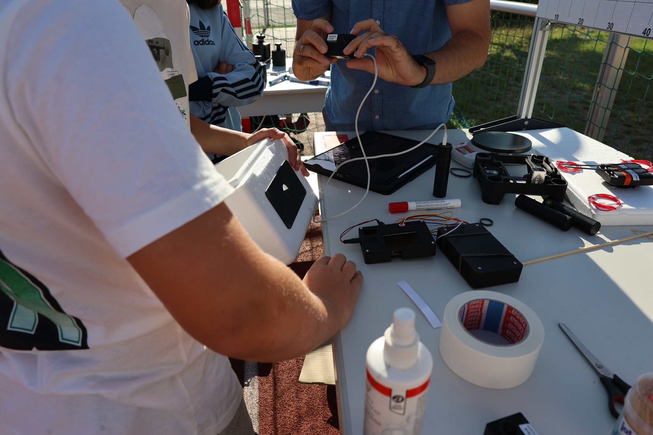 Personen arbeiten an einem Tisch mit Elektronikkomponenten und weißem Schaumstoffblock.
